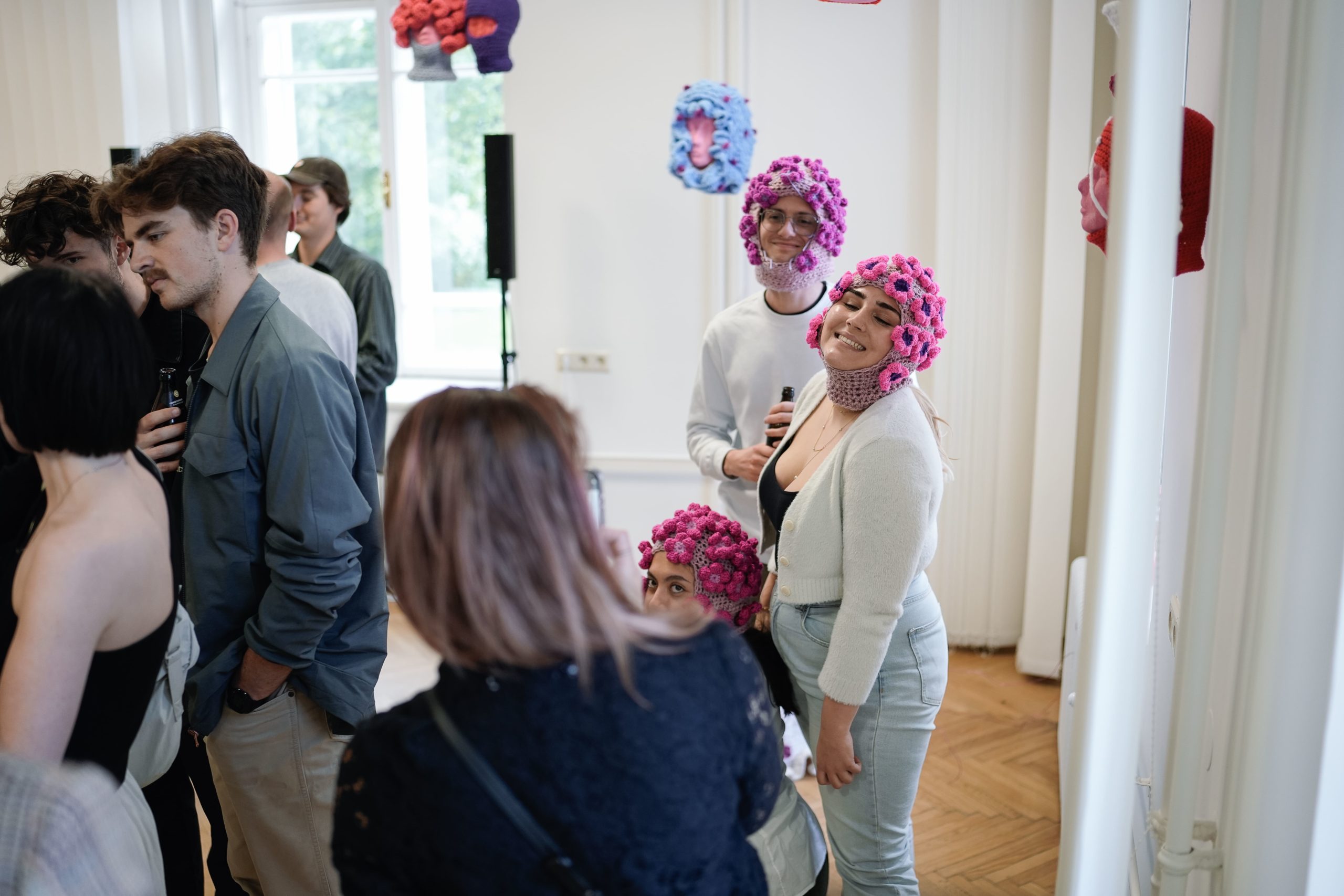 Zu sehen sind Besucherinnen und Besucher der Ausstellung. Auf der linken Seite unterhalten sich einige miteinander, auf der rechten Seite macht eine Besucherin ein Foto von drei Menschen, die Balaclavas mit aufgenähten Häkelblumen in verschiedenen Farben auf haben. Im Hintergrund hängen Styropor-Köpfe mit weiteren Balaclavas: eine graue mit hellroten gehäkelten Kugeln neben einer violetten, eine hellblaue mit lila Punkten und etwas verdeckt an der rechten Wand eine rote mit einer weißen Schnur über dem Gesicht des Styropor-kopfs.