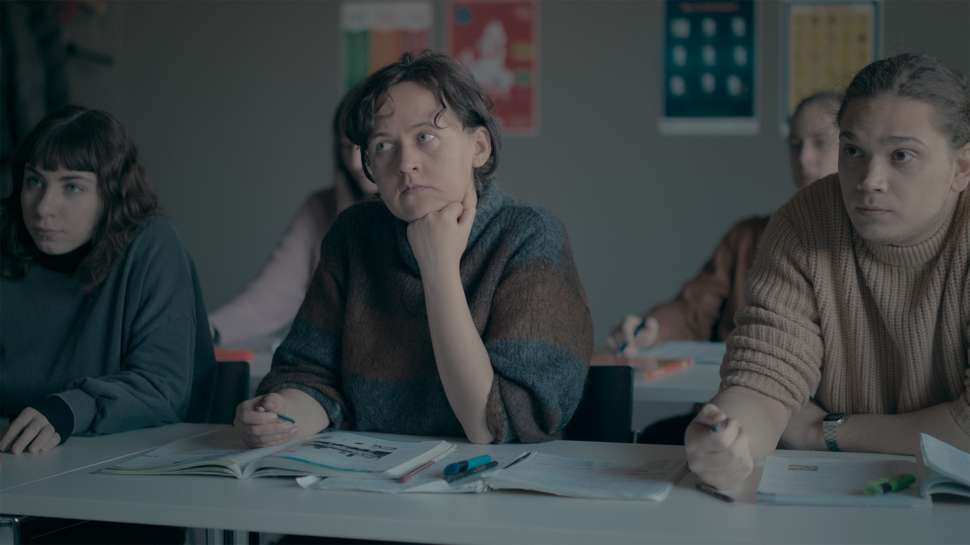 The main character is sitting by the desk in the class among other students.