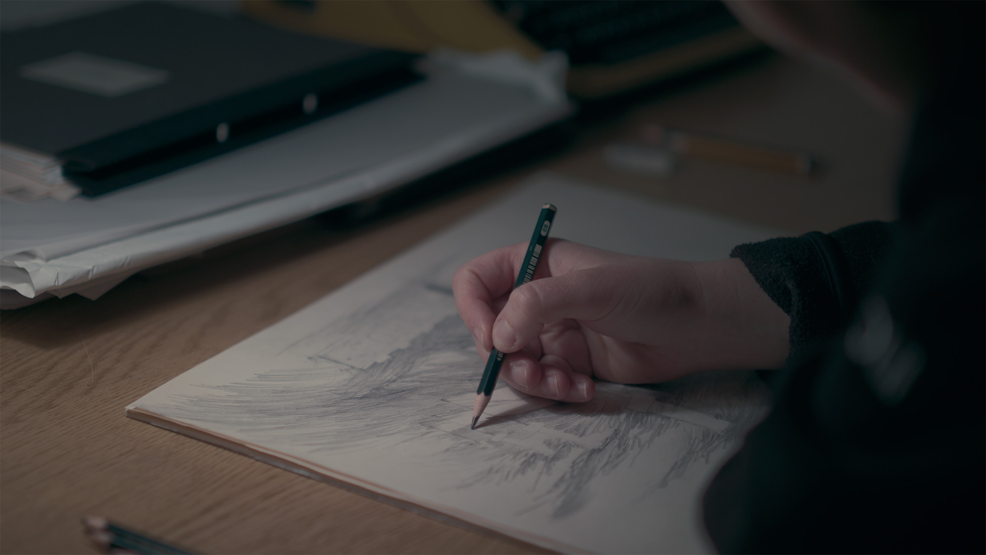 Close up of the hand with a pencil, drawing a graphic image on a white sheet of paper on a desk. The drawing is not clear.