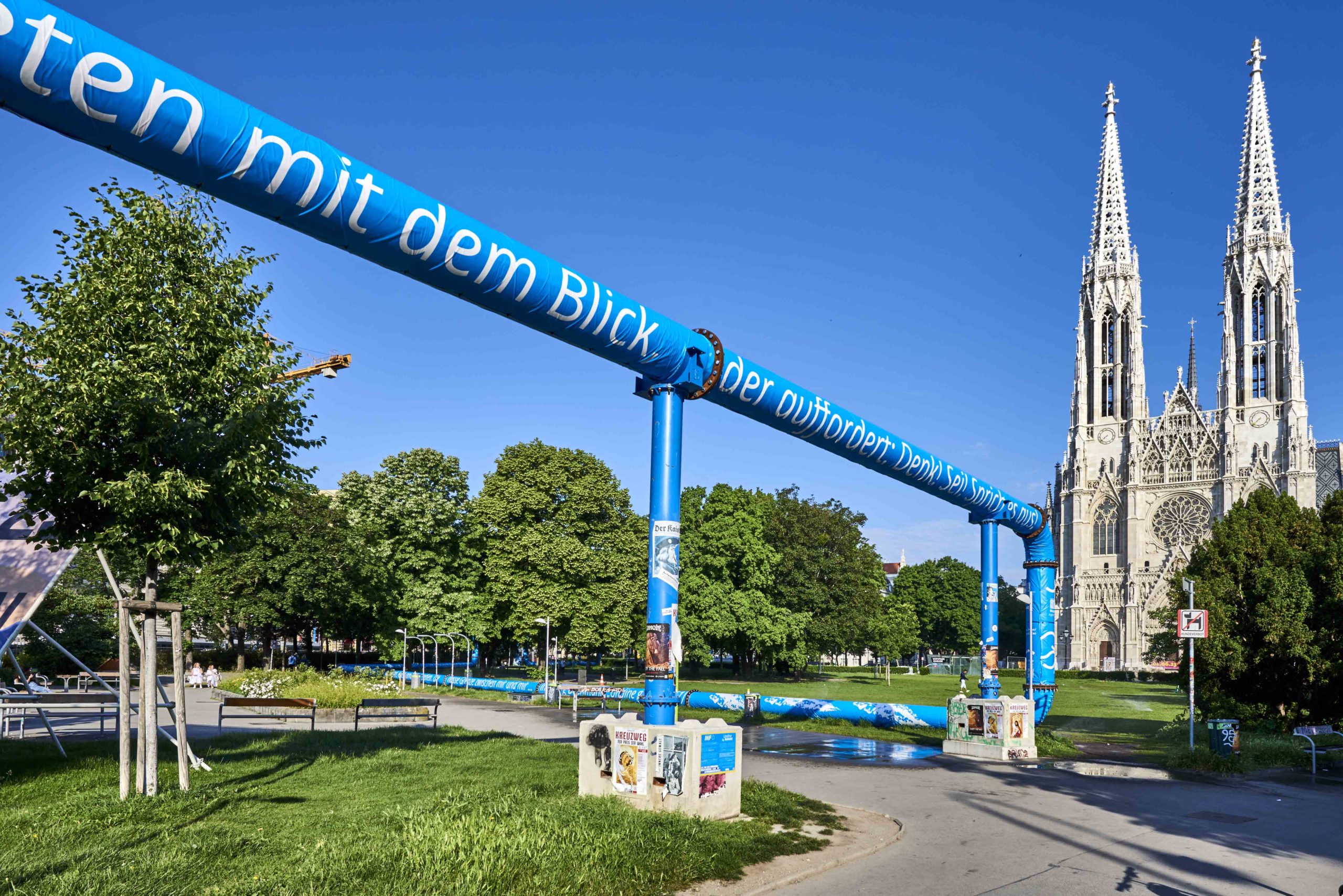 Man kann einen Ausschnitt der Rohre mit den blauen Planen sehen. Darauf sind Zitate von Ingeborg Bachmanns „Undine geht“ gedruckt. Der Himmel ist strahlend blau, auf der rechten Seite sieht man einen Teil der Votivkirche.