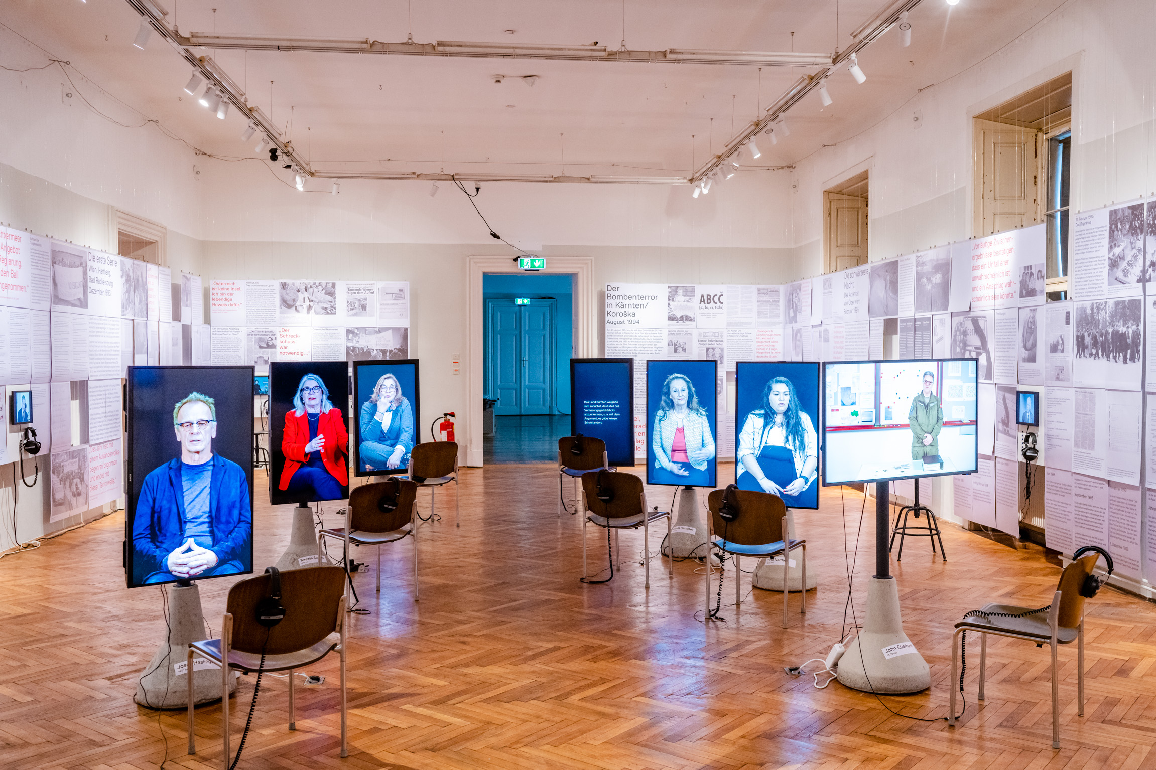 Ausstellungsansicht im Volkskundemuseum Wien. Zu sehen sind sieben der insgesamt neun Videointerviews. V.l. Josef Haslinger (Schriftsteller), Terezija Stoisits (ehm. Politikerin), Silvana Meixner (ehem. Moderatorin ORF-Minderheitenredaktion), Joža Messner (Mitinitiator d. zweisprachigen öffentlichen Volksschule, Klagenfurt), Ursula Hemetek (Mitbegründerin Initiative Minderheiten), Tina Nardai (Journalistin aus Oberwarter) und John Eberhardt (Entschärfungsdienst Innenministerium). Weitere Interviewpartner*innen sind nicht im Bild, sondern im Eingangbereich der Ausstellung zu sehen: Andreas Peham (Rechtsextremismusexperte, DÖW) und Armin Thurnher (Falter Herausgeber). An den Wänden hängen Zeitungs- und ORF-Fernseh- und Hörfunkberichte, Fotos und Dokumente.