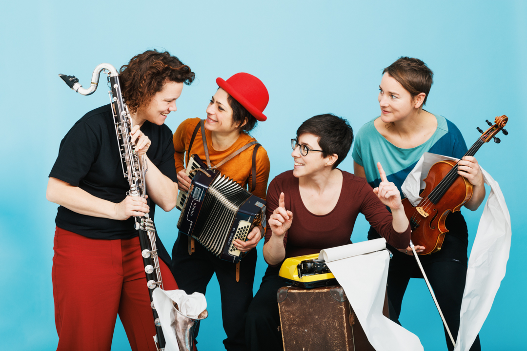 Vor fröhlich blauem Hintergrund ist das gesamte Produktionsteam. Julia Schreitl hält die Bassklarinette und schaut Johanna Jonasch, die Regisseurin, die an der gelben Schreibmaschine sitzt, an. Franziska Adensamer trägt einen roten Hut und spielt das Akkordeon. Judith Reiter hält Bratsche und Bogen in der Hand.