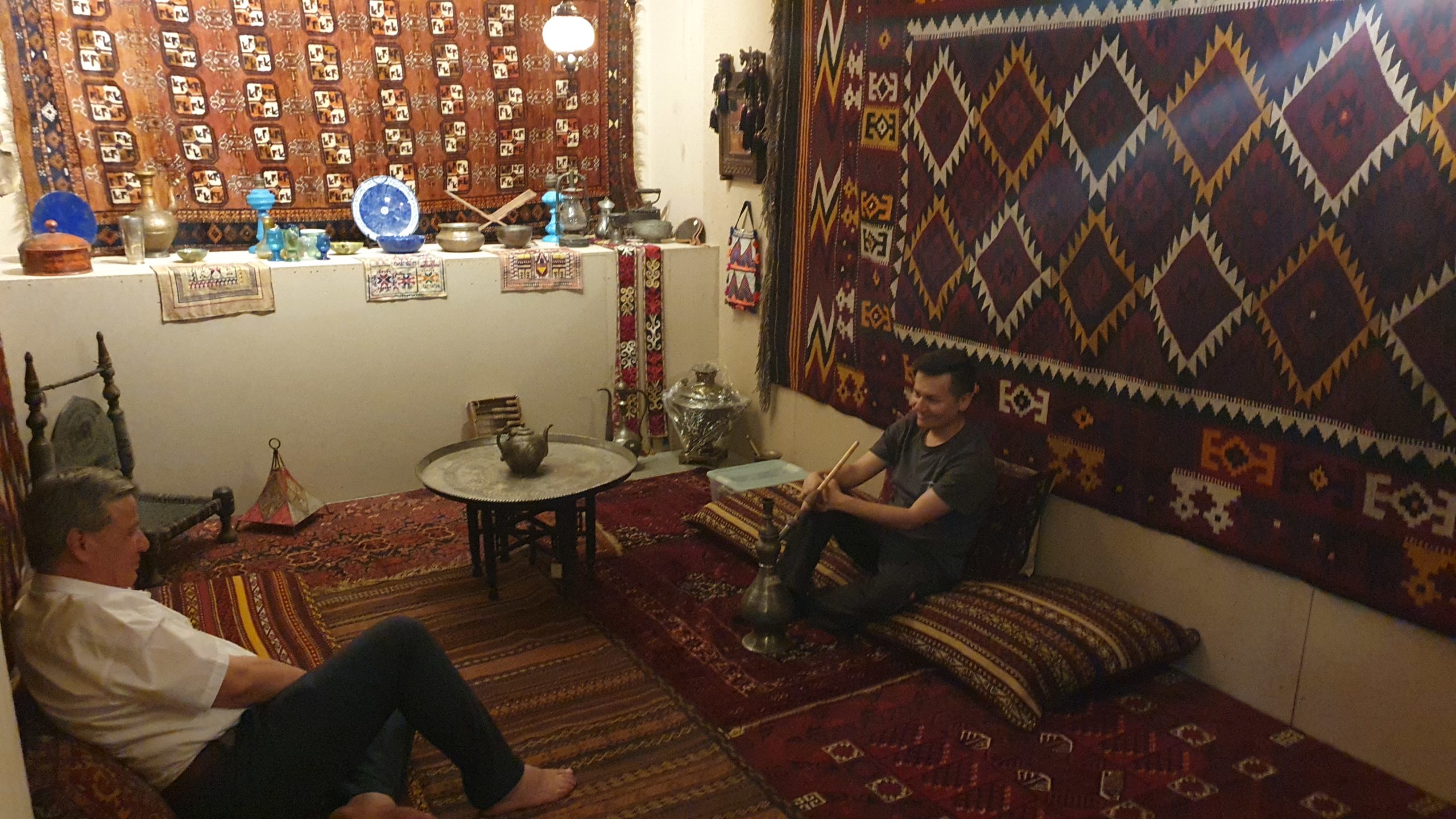 Das zweite Bild zeigt die Kunstinstallation von Innen. Dabei wird der Raum des Hauses dargestellt, der mit afghanischen Teppichen und Interior ausgekleidet ist. An den Wänden hängen ebenso traditionelle bunte Teppiche. In der Mitte des Raumes steht ein kleiner runder Tisch mit einer Teekanne drauf, um den zwei Männer auf gemütlichen Pölstern sitzen. Im Hintergrund sieht man einen kleinen Vorsprung, auf dem weitere afghanische Behältnisse und Vasen stehen.