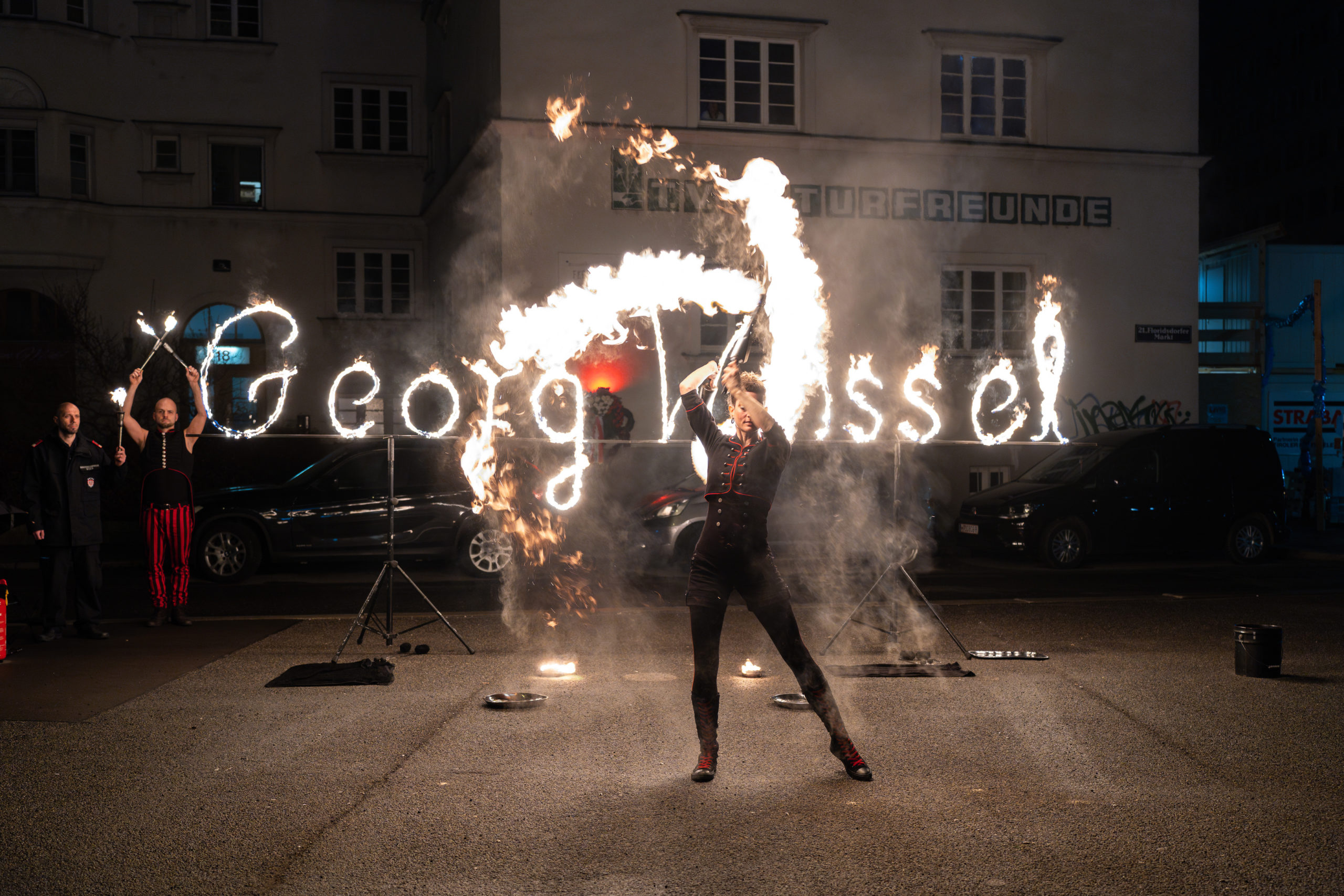 Schlussszene mit Flame Rain Theater: Frau performt mit brennenden Fackeln, im HIntergrund der aus brennenden Buchstaben gebildete Namen Georg Weissel