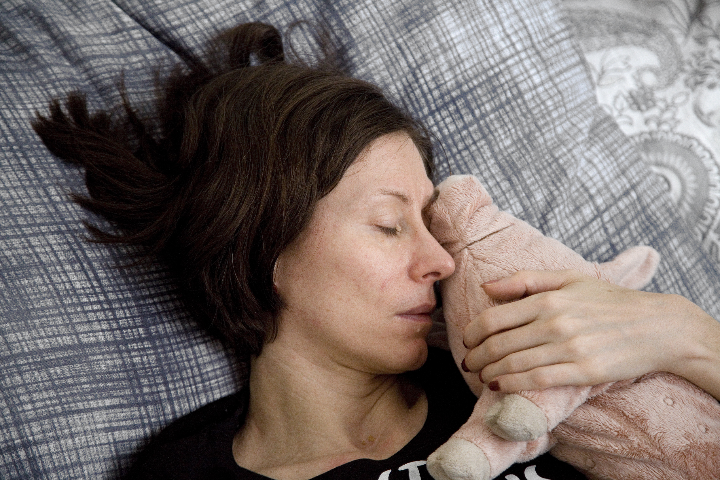 Coverfoto der Ausstellung Crash! Das Foto zeigt das Porträt von Judith Schoßböck (Black Ferk Studio), in ihrem Bett auf einem grau-weißen Kopfpolster liegend. Ihr Gesicht ist von der Seite zu sehen, ihre Augen sind geschlossen. In ihrer linken Hand hält sie ein rosa Stoffschwein an ihr Gesicht.