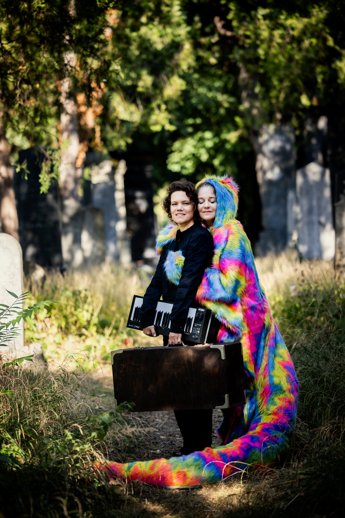 Man sieht die zwei Performerinnen auf einem Friedhof. Eine trägt schwarze Kleidung und hält ein Keyboard sowie einen Koffer in ihren Händen. Sie wird von der zweiten Performerin, die ein regenbogenfarbenes Ganzkörper - Kuschelkostüm mit einem langen Schweif trägt umarmt.