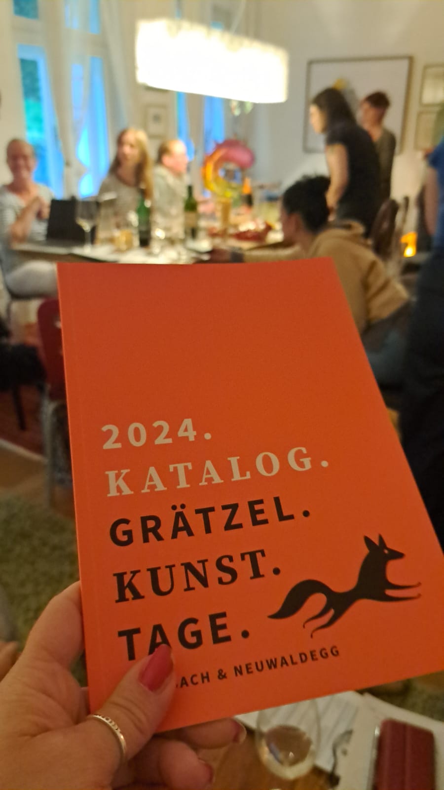 Man sieht in Vordergrund den orange farbigen Kunstkatalog der ersten Grätzel.Kunst.Tage mit unserem Logo, einem schwarzen Fuchs. In ihm sind alle Kunstschaffenden, die an der Konzeption der Grätzel.Kunst.Tage mitgewirkt haben, mit ihren Lebensläufen und einem ihrer Werke zu finden. Im Hintergrund sieht man das ehrenamtliche Team bei der Arbeit, bei einem unserer insgesamt acht Projekttreffen zur Planung und Durchführung der Grätzel.Kunst.Tage.