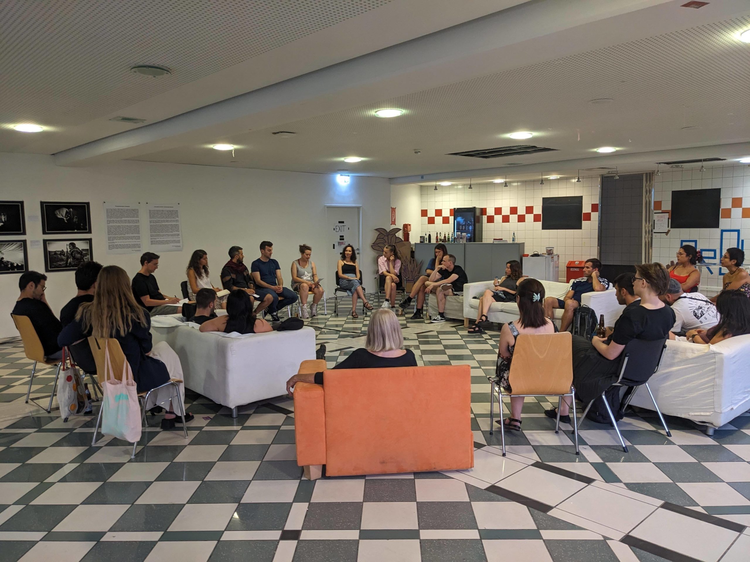 Eine Gruppe von Personen sitzt im Kreis auf Sofas und Stühlen und hört einer Diskussion zu, im Hintergrund befinden sich Bilder von einer Ausstellung.