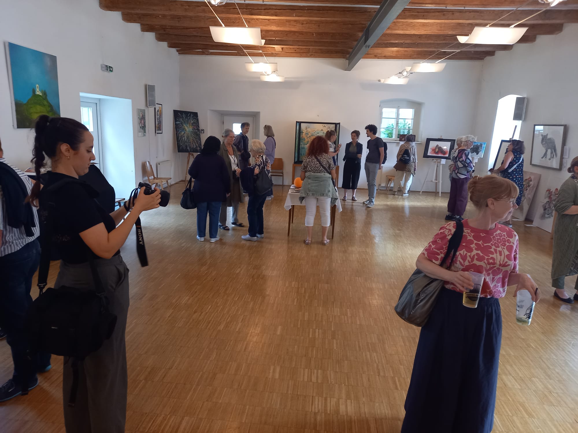 Auf dem Bild sieht man den Pfarrsaal in dem die Werke der Kunstschaffenden teilweise auf Staffeleien oder hängend präsentiert wurden, Besucher:innen, die sich die Werke ansehen, teilweise im Gespräch mit den Künstler:innen.