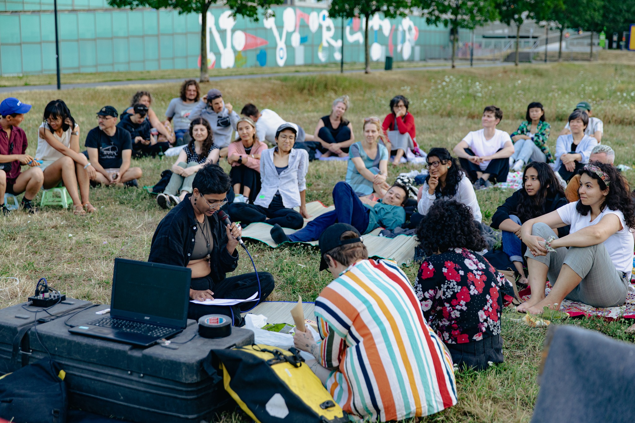 Lesung von Manu Sharma und Anushri Rastogi in einer Wiese des Donauparks. Es ist Dämmerung, viele Menschen sitzen auf Picknickdecken und hören aufmerksam zu.