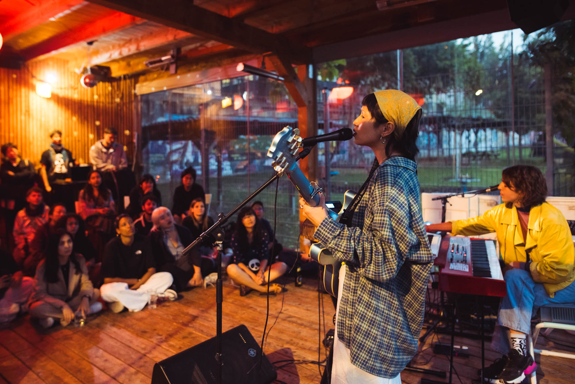 Konzert von SAKURA bei der Auftaktveranstaltung in Central Garden. SAKURA spielt Guitarre und singt, das Publikum sitzt am Boden und schaut gebannt zu.