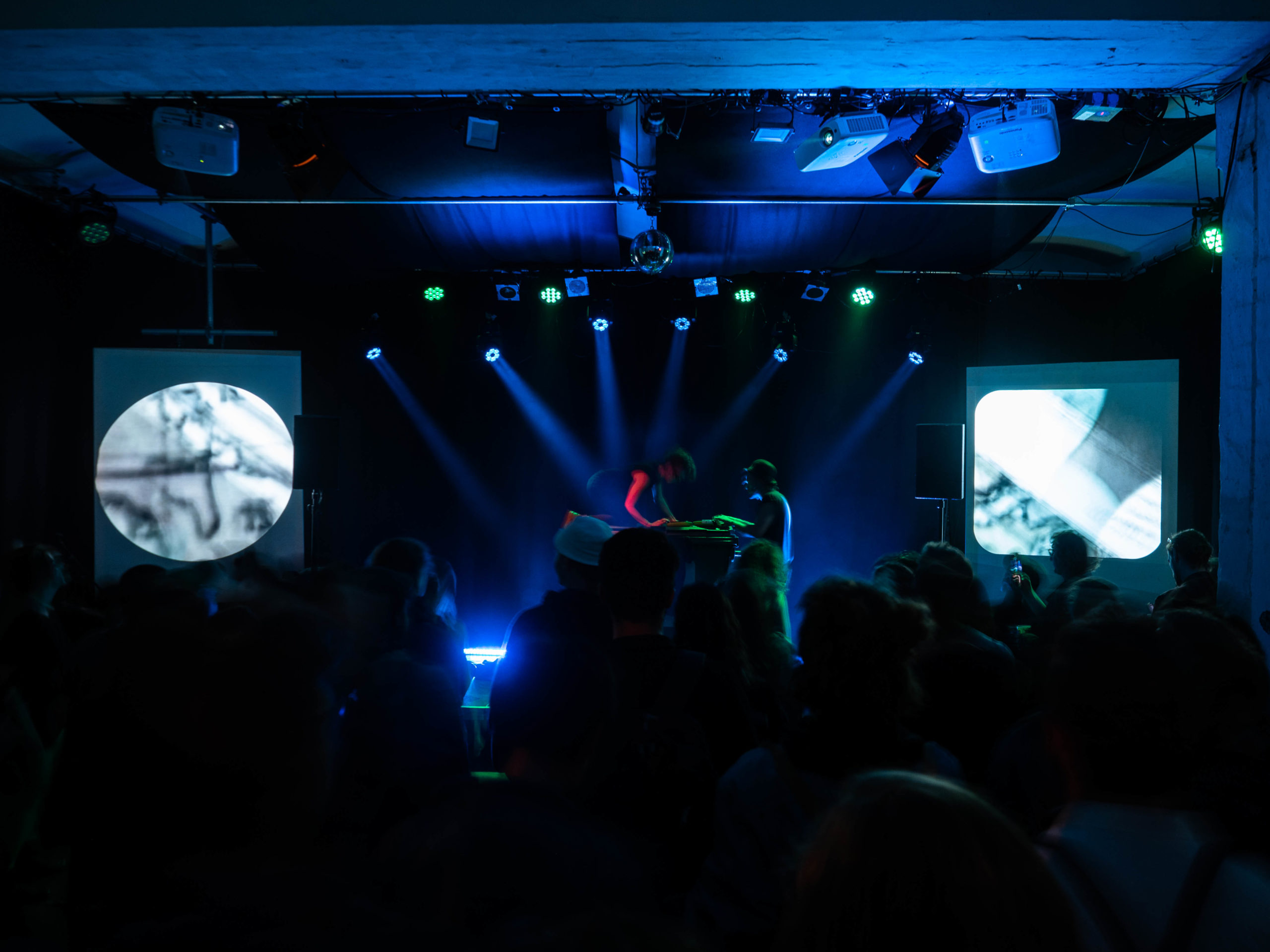 Der Veranstaltungsraum in der Alten Schmiede, hier während einem Konzert zu sehen. Man sieht dunkelblaues Licht, Nebel, dunkle Menschenshilouetten, im Hintergrund die Bühne.