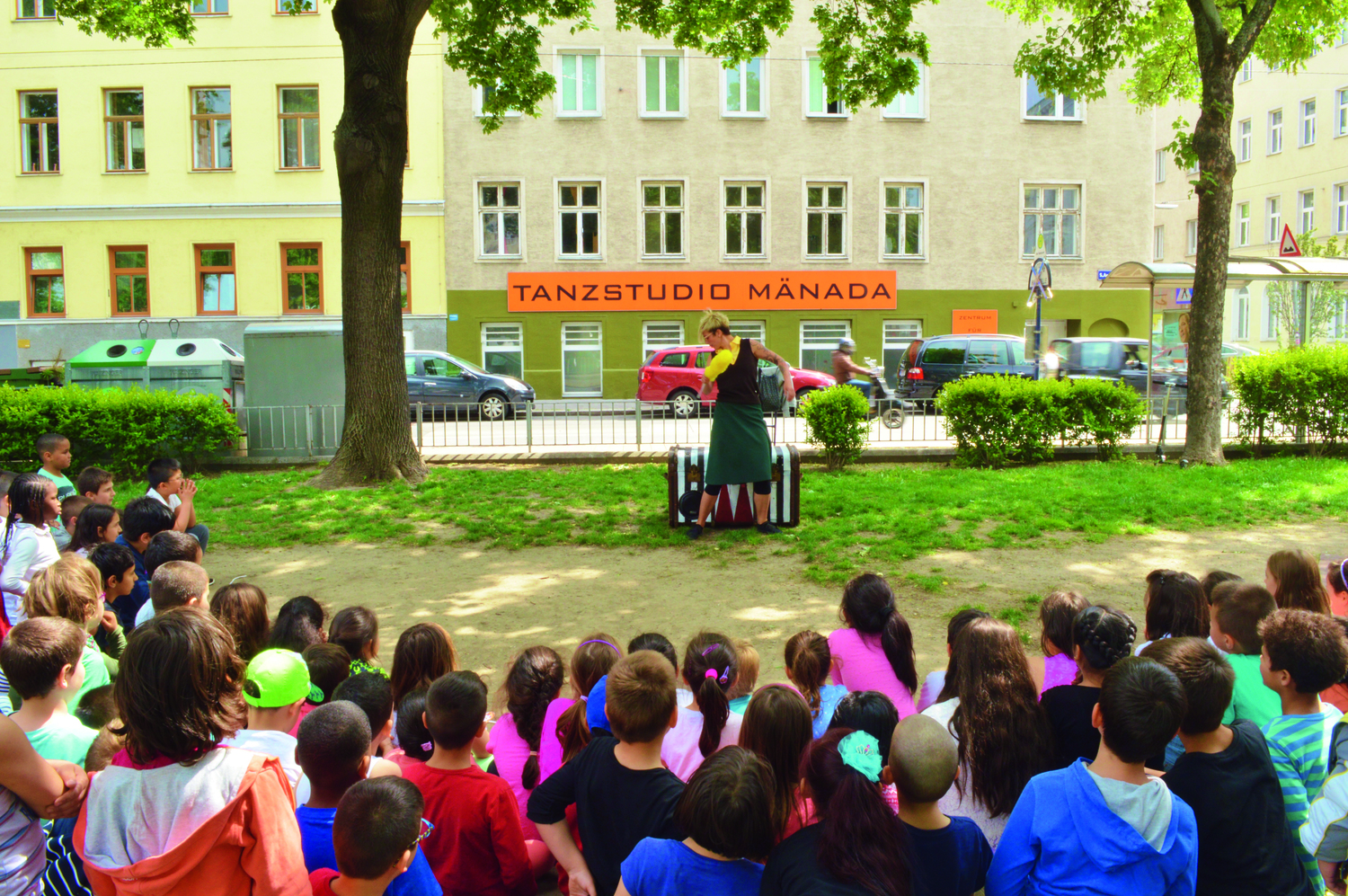 Ein innerstädtischer Wiesenplatz, Kinder schauen auf die Grünfläche, auf der man* eine Person mit einer Kiste sieht