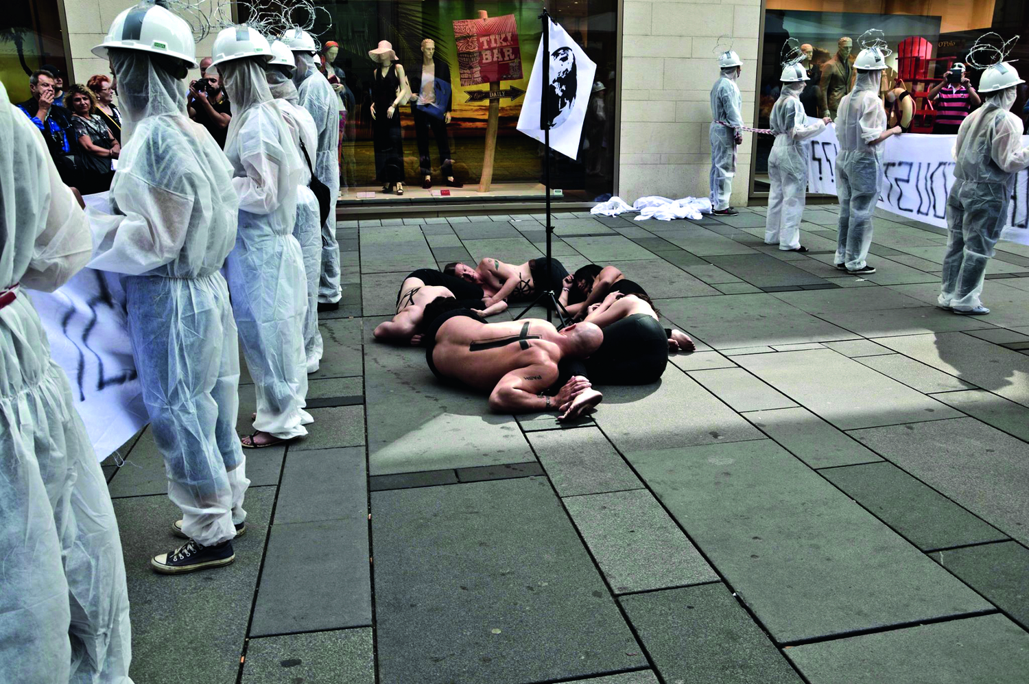 Foto einer Straßenaktion, an den Seiten stehen Menschen in weißen Hygieneoveralls, in der Mitte liegen 5 Personen in schwarzer Unterwäsche zusammengekümt auf dem Asphalt