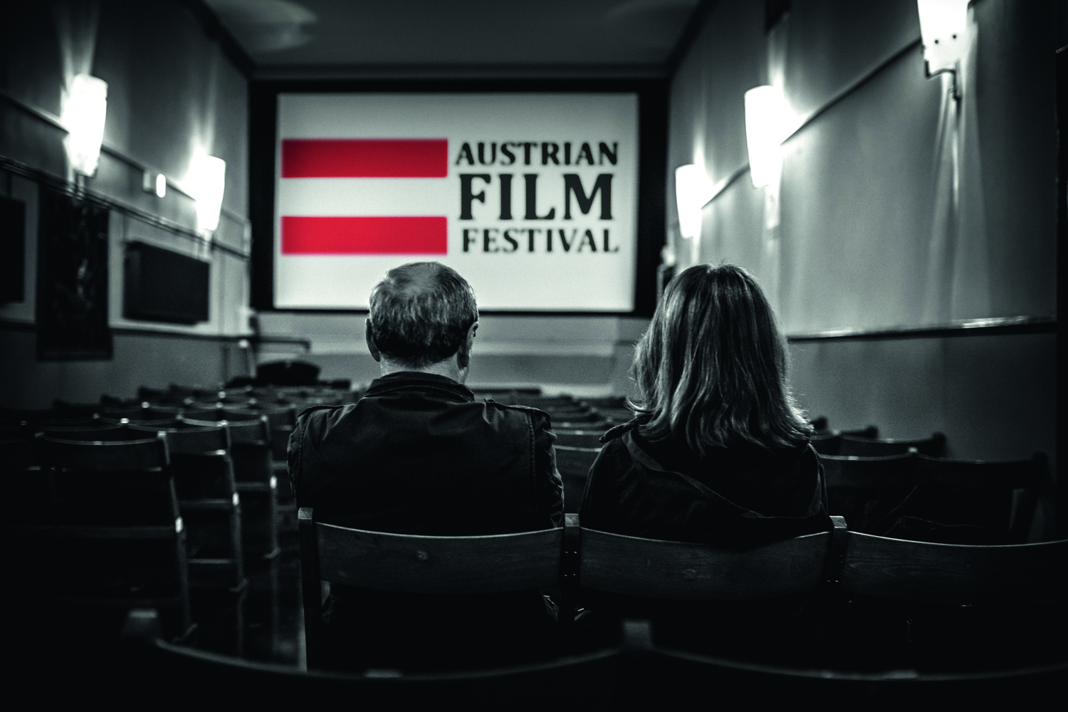 Schwarzweißfoto, 2 Menschen sitzen in einem Kino (BSL) und schauen auf die Leinwand, dort sieht man das Logo des Festivals, wobei die österreichische Fahne daneben als einziges in Farbe zu sehen ist 