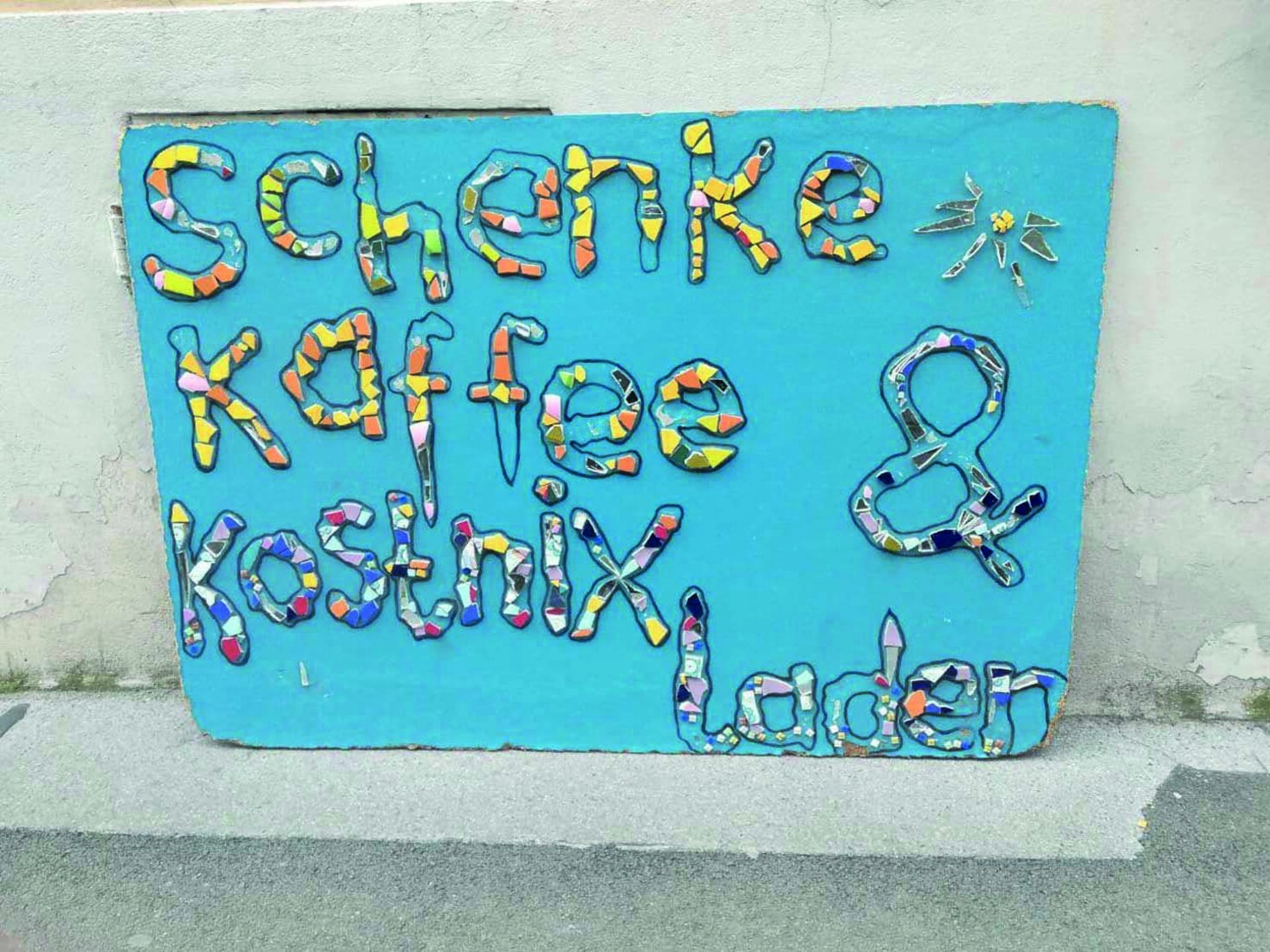 Selbstgemachtes Plakat, auf blauem Hintergrund stehen in bunten Buchstaben "Schenke Kaffee" und "Kostnix-Laden"