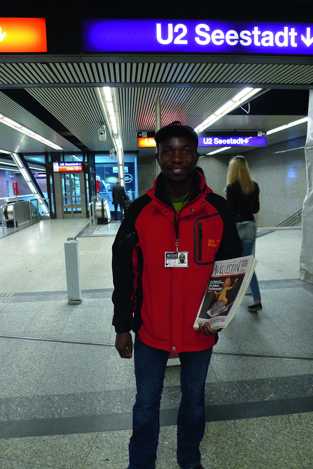 Porträtfoto eines Mannes, den man an einer U-Bahn-Station stehen sieht, er hält die Straßenzeitung Augustin in der Hand und lächelt nach vorne, den Betrachter:innen entgegen