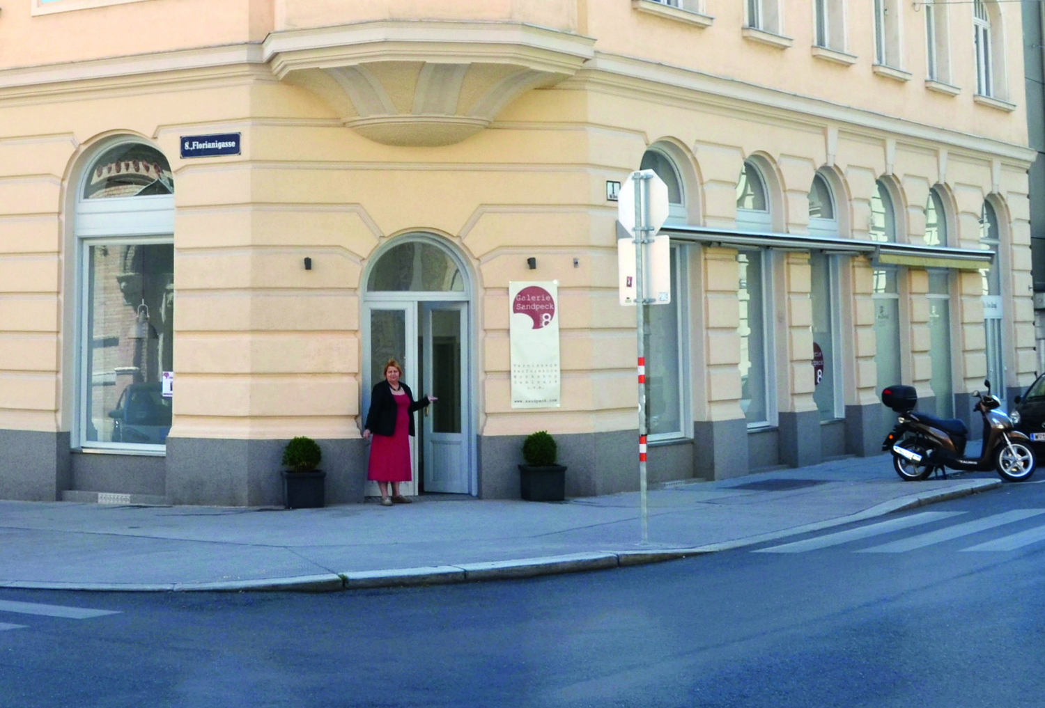 Man sieht eine Straßenecke bzw. (fotografiert vom anderen Ende aus) ein Eckhaus, Gründerzeit, gelb, an der Eingangstür zum am Eck befindlichen Straßenlokal steht eine Frau, sie zeigt mit der flachen Hand auf die geöffnete Tür und lacht nach vorne Richtung Betrachter:innen