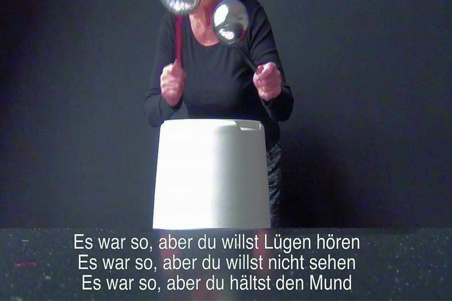 Eine Frau, deren Körper ab der Schulter nicht zu sehen, ist, steht in einem grauen Raum und trommelt mit zwei Suppenschöpfern auf einen verkehrt auf den Boden gestellten Plastikkübel, darunter stehen drei Zeilen Text