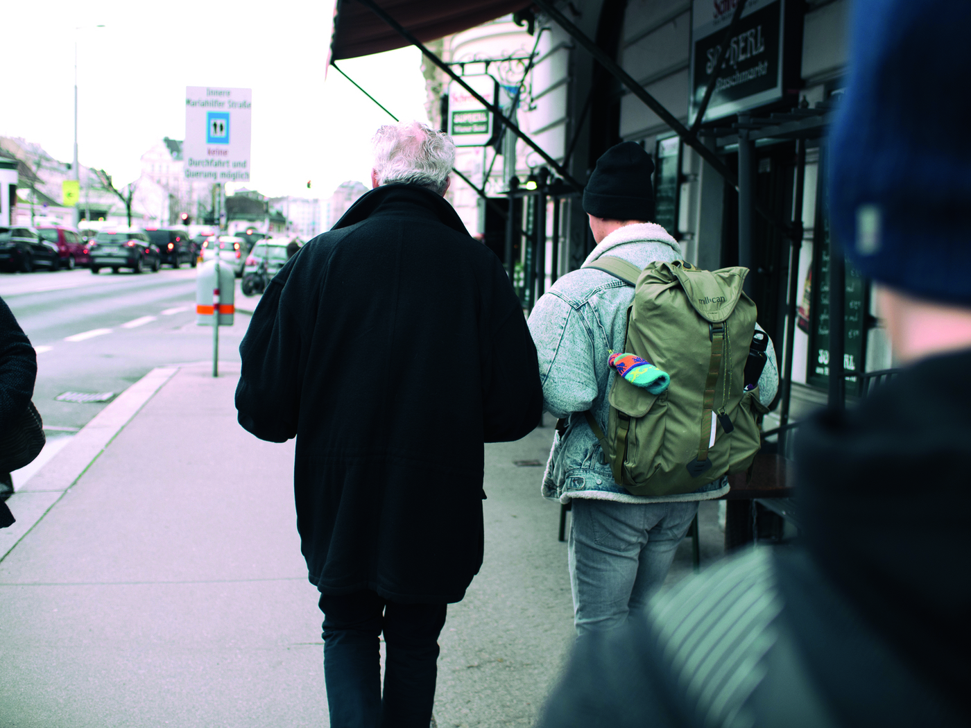 Zwei Personen gehen auf der Straße, man sieht sie nur von hinten, die linke Person hat weiße Haare, die rechte eine schwarze Wollmütze und trägt einen Rucksack