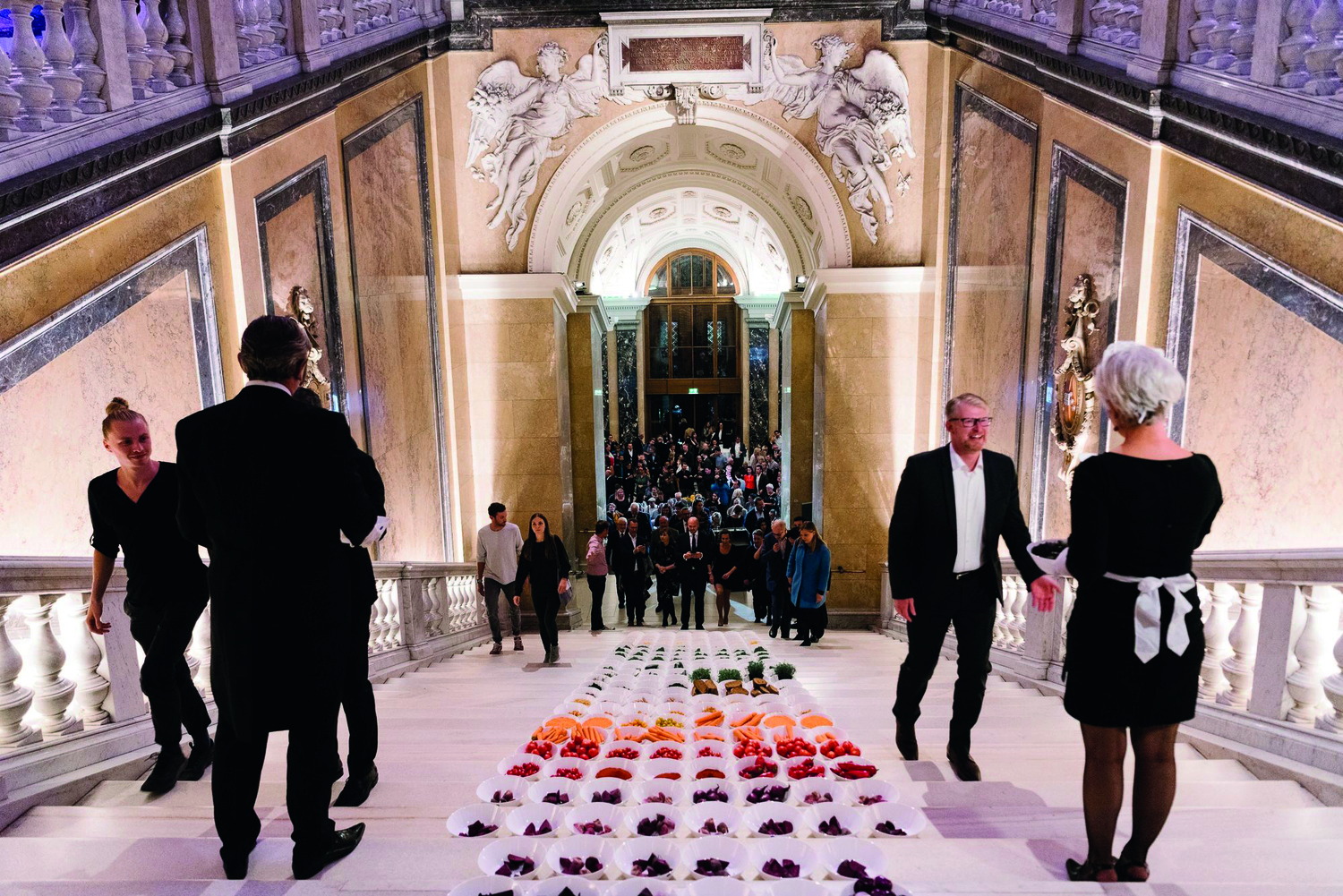 Stufenaufgang in einem Palais, Menschen gehen die Treppen hinauf, auf denen kleine Schüsseln mit Essen stehen, daneben "Kellner:innen", die das Publikum empfangen