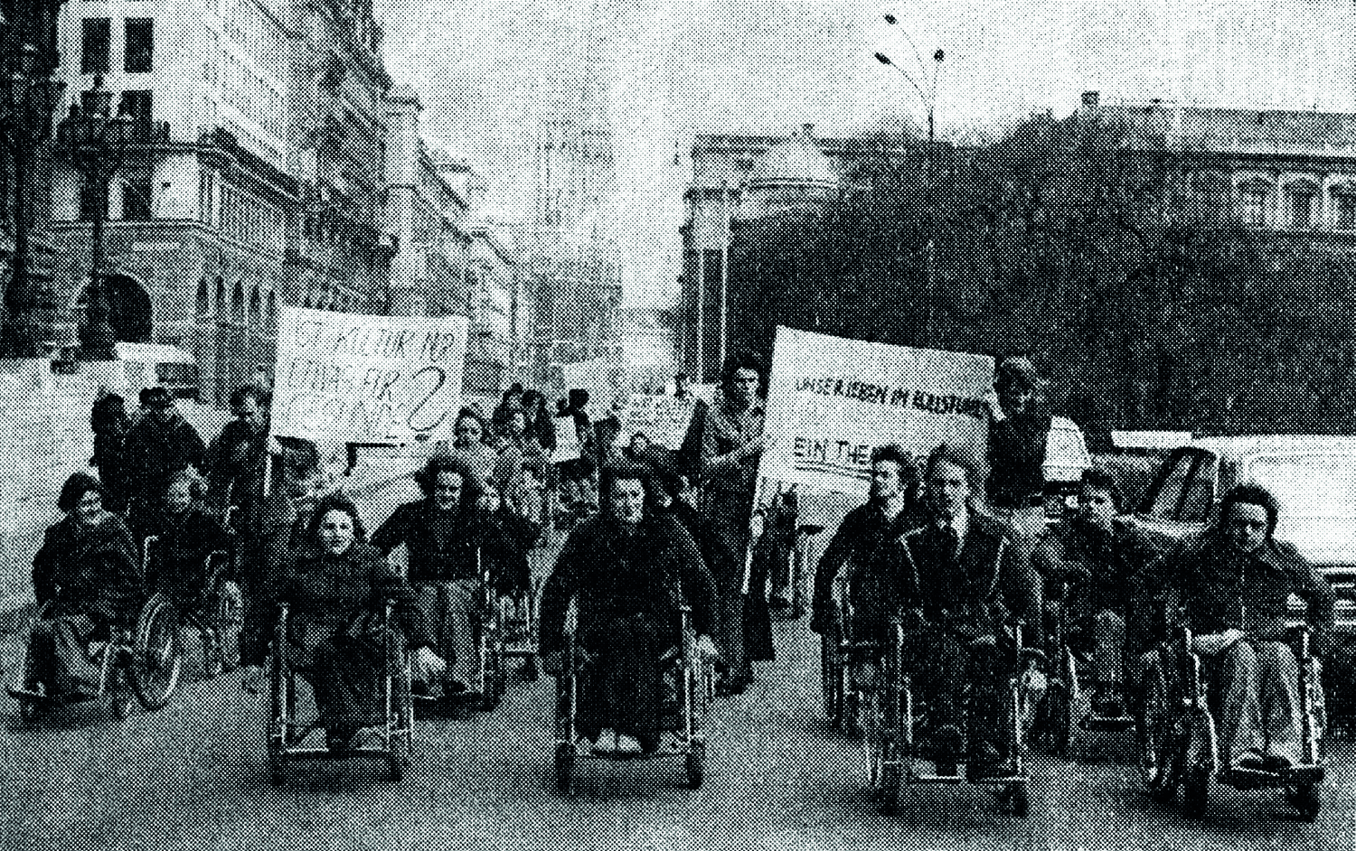 Historisches Schwarzweißbild, Rollstuhlfahrer:innen auf der Straße rollen, teils mit großen Plakaten, in Richtung Kamera (Protest im Rollstuhl ("Kurier", März 1974) 