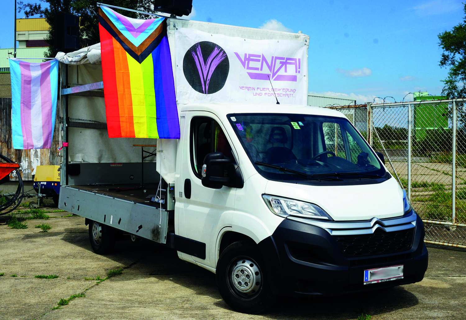 Ein Foto des Venga-Busses, ein weißer Planen-LKW, die an einer Seite offene Plane hinten hat Regenbogenfahnen und das Logo des Projekts