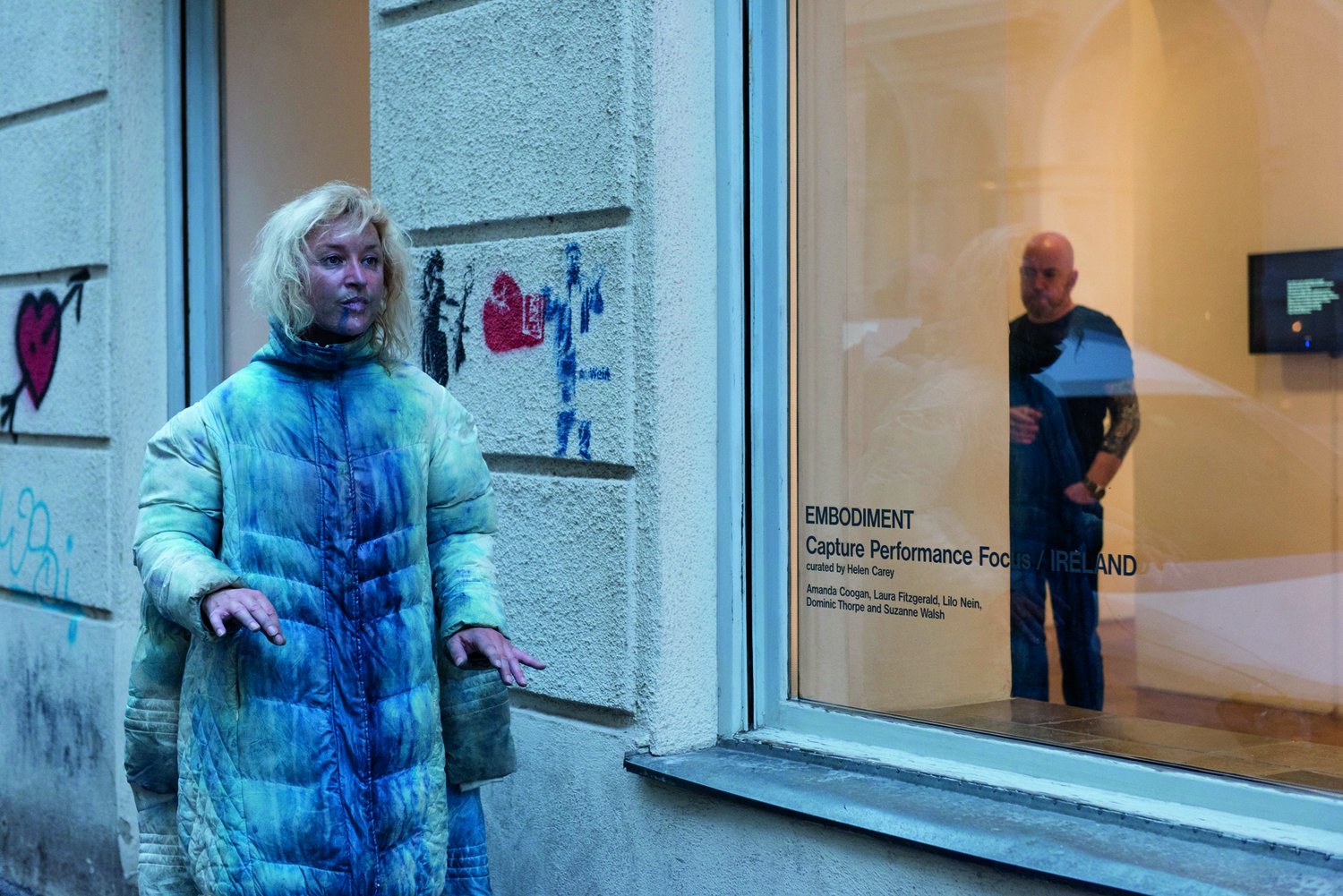 Der Ausstellungsraum von außen, drinnen ist es hell, Bilder hängen an den Wänden, eine Person steht im Raum; vor der Galerie geht eine Frau* in einem weißen Mantel, der blau eingefärbt wurde