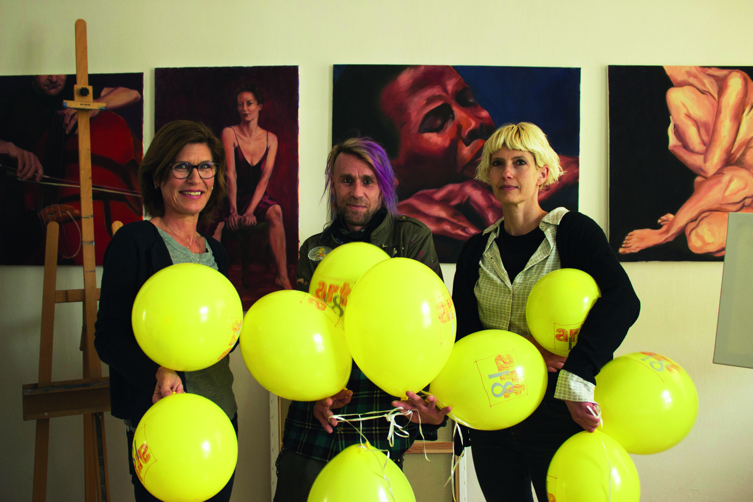 Drei Personen stehen mit gelben Ballons in der Hand in einem Galerieraum vor einer Bilderwand