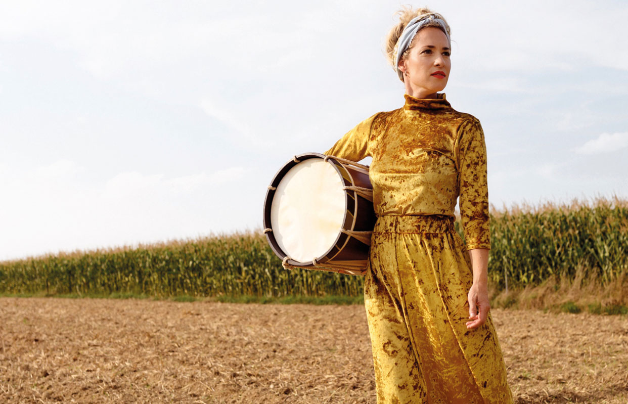 Künstler:innen-Porträt, die Sängerin steht mit einer Trommel unter ihrem rechten Arm und in einem goldenen Kleid auf einem trockenen Feld, hinter ihr ein Maisfeld