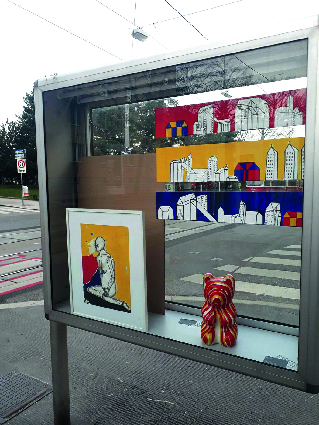 Eine Glasvitrine im öffentlichen Raum, in der sich Bilder in Gelb-Rot-Blau sowie eine Bärenskulptur in denselben Farben befinden