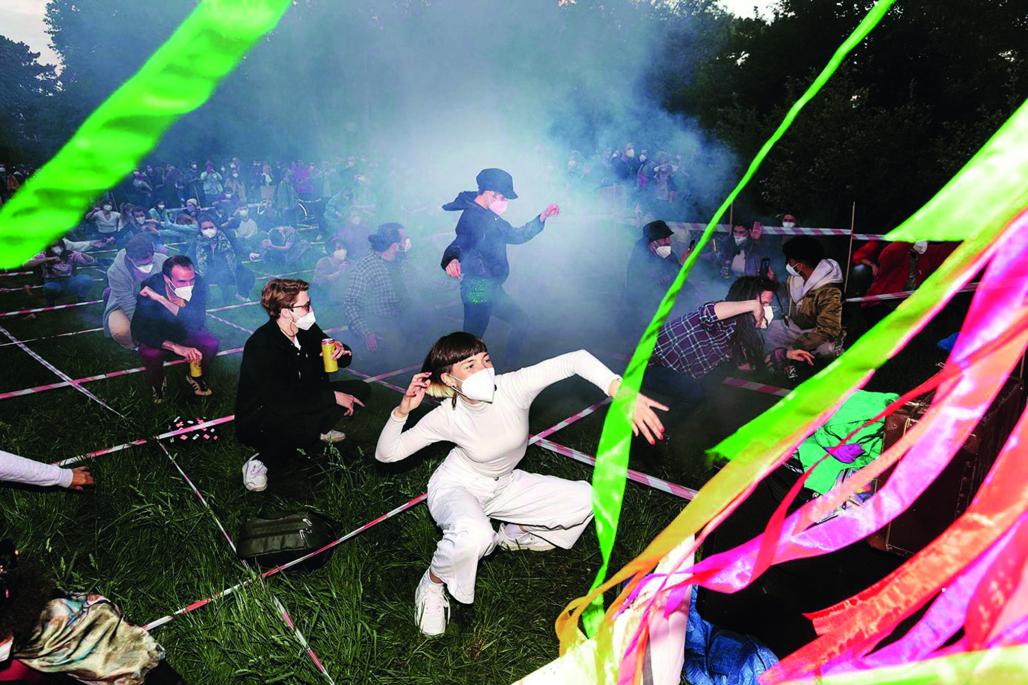 Foto eines der Raves, Personen knien im Gras, rechts vorne sieht man bunte Plastikbänder, die sich im Wind zu bewegen scheinen