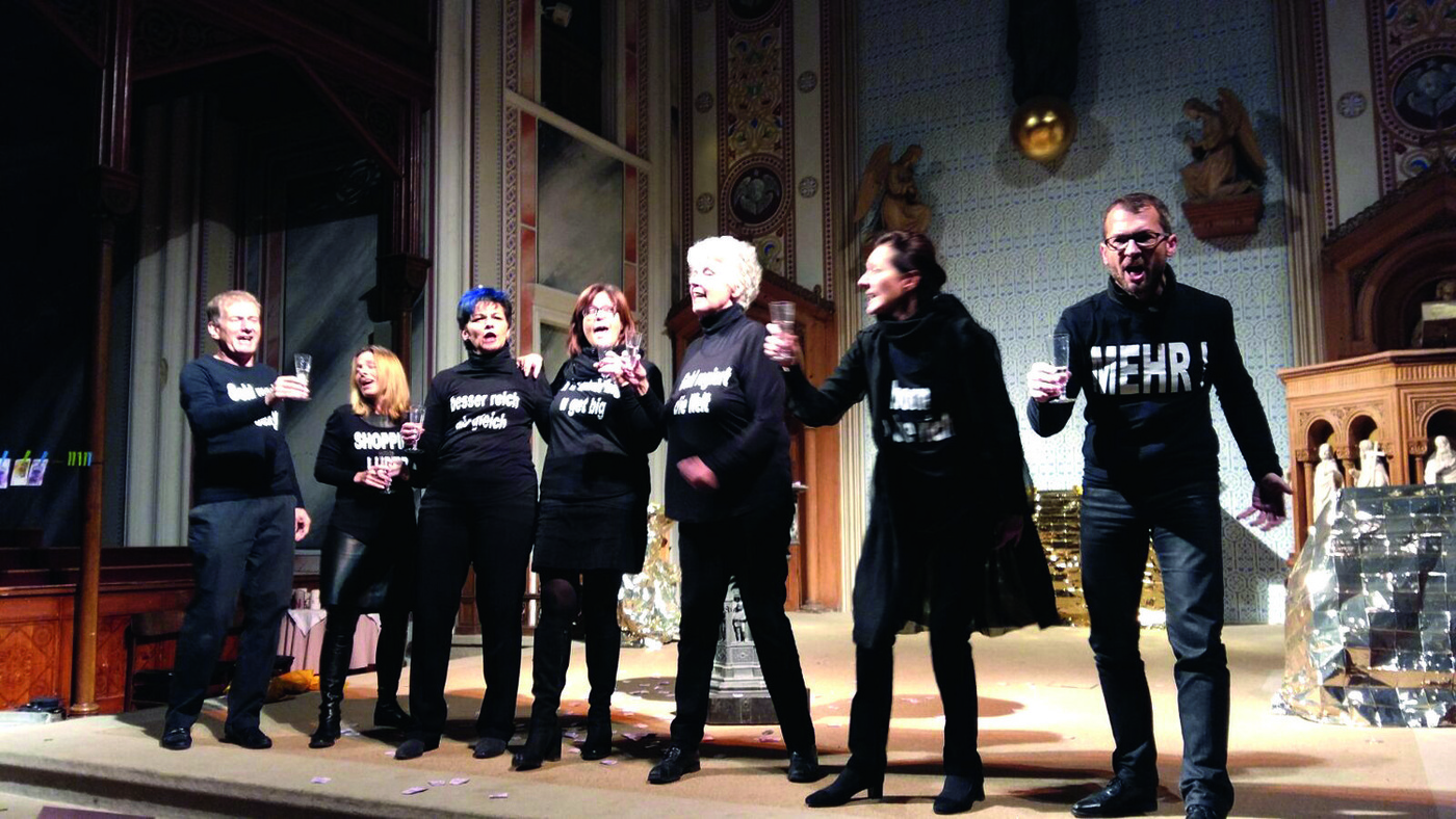 Szenenfoto, in einem Raum, der an eine barocke Kirche oder ein Palais erinnert, stehen die Darsteller:innen nebeneinander, sie tragen schwarze T-Shirts mit Schriftzügen, halten Sektgläser in den Händen und scheinen einander zuzuprosten oder auch zu singen