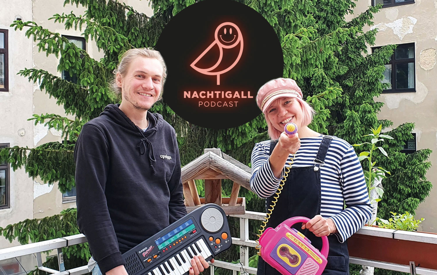 Maria Danilina und Sandro Nicolussi im Porträtfoto, auf einem Balkon, mit Plastikinstrumenten und einem bunten Mikro in der Hand und lachend nach vorne blickend