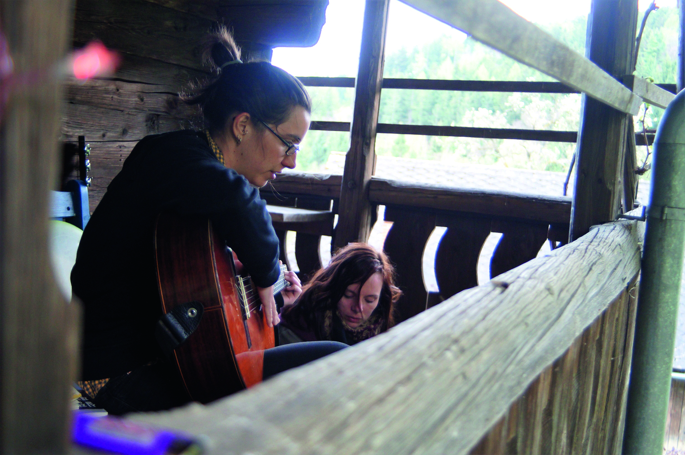 Eine Art Holzbalkon, in der 2 Frauen sind, eine spielt Gitarre, die andere sitzt oder kniet hinter ihr