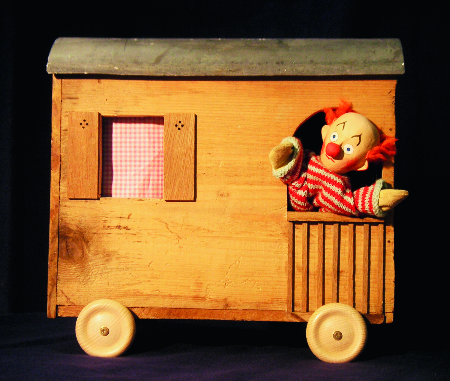 Aus einem kleinen, hellen Holzeisenbahnwagon schaut ein Puppen-Clown heraus