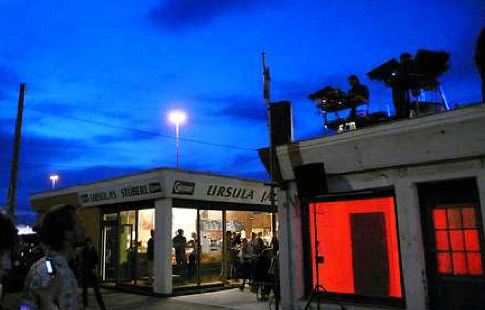 Ein Markt bei Nacht, zwei Markthütten sind innen hell erleuchtet, eine ganz in Rot, der Himmel ist in strahlen dunkles Nachtlicht getaucht