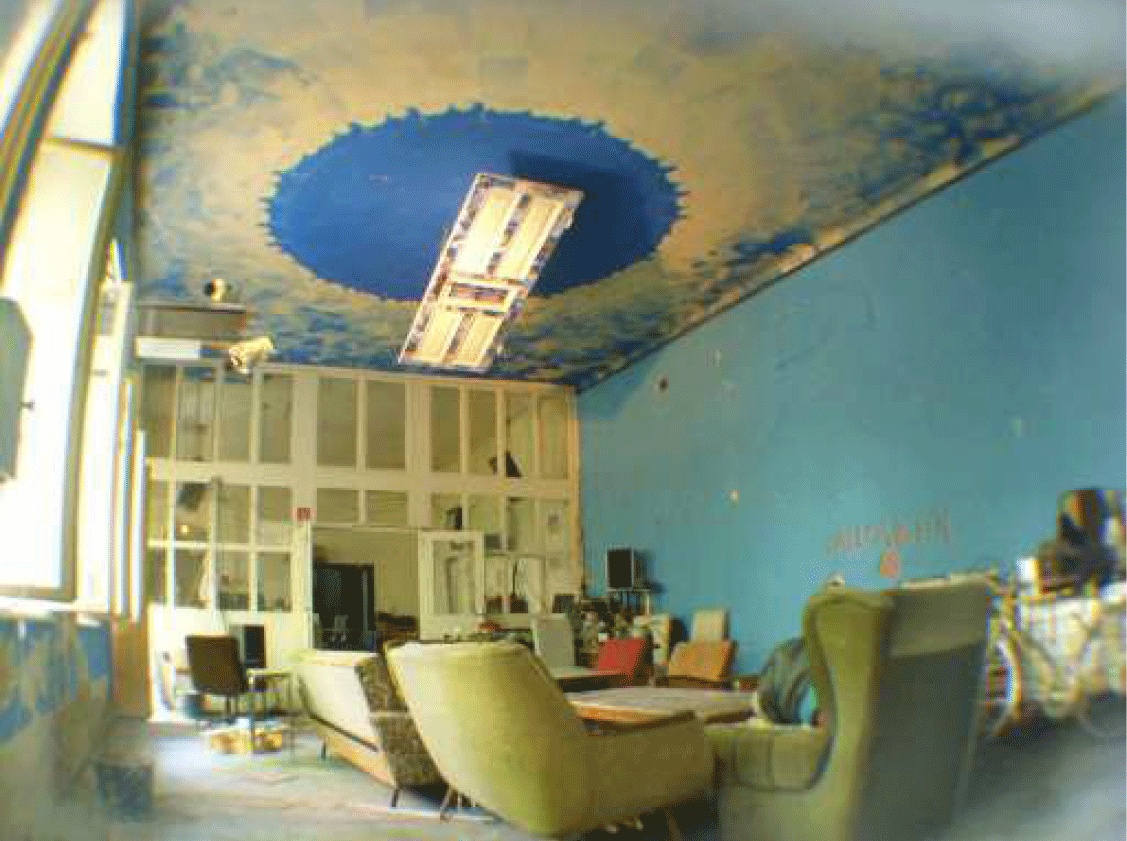 Eine gemütliches Wohnzimmer-Ambiente, hellblaue Wand, links eine Fensterfront, die Decke ist shabby weiß mit einem strahlend blauen Kreis in der Mitte, die hintere Wand ist eine alte, wandbreite Holztüre mit Fenster, im Raum stehen gemütliche alte Fauteuils, Stühle und Sofas