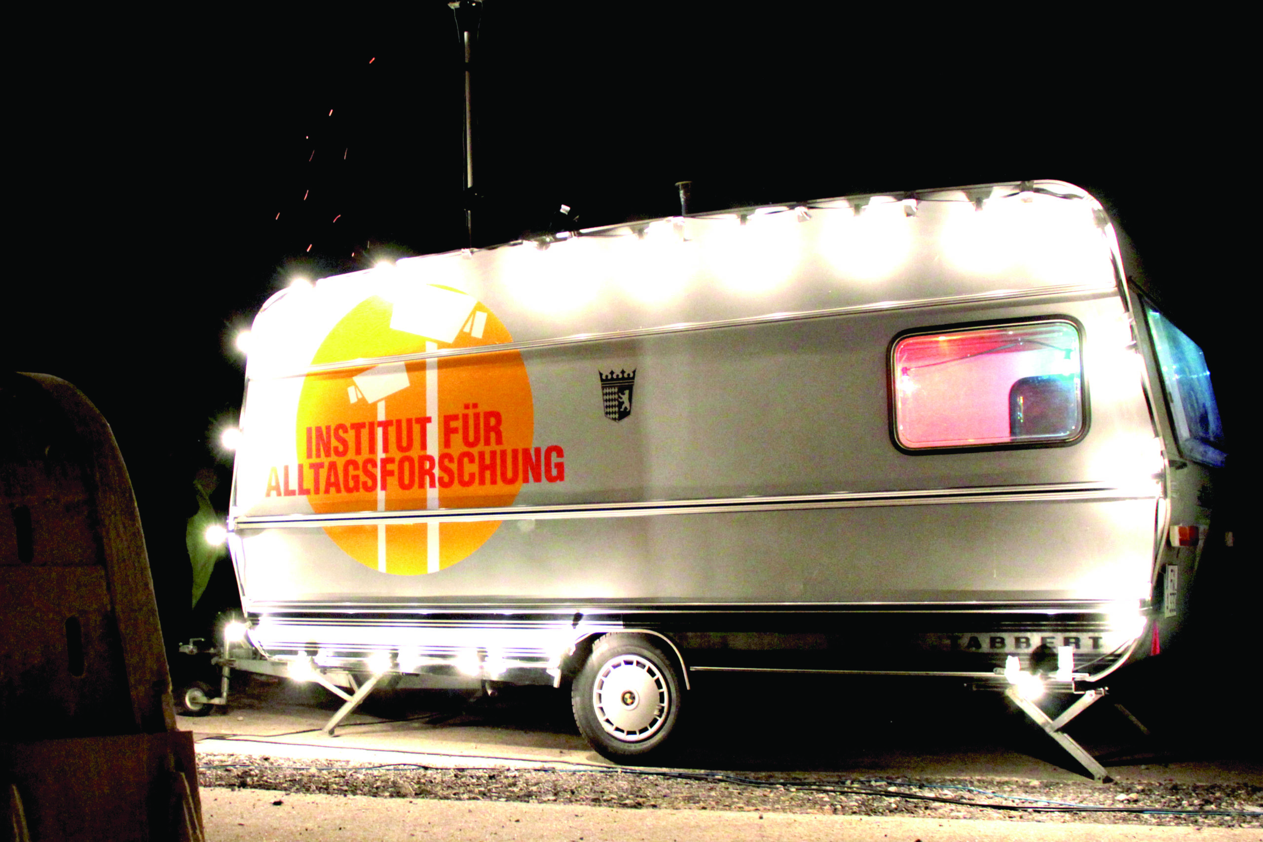 Ein weißer, innen erleuchteter Campingwagen bei Nacht mit dem Logo des Projekts