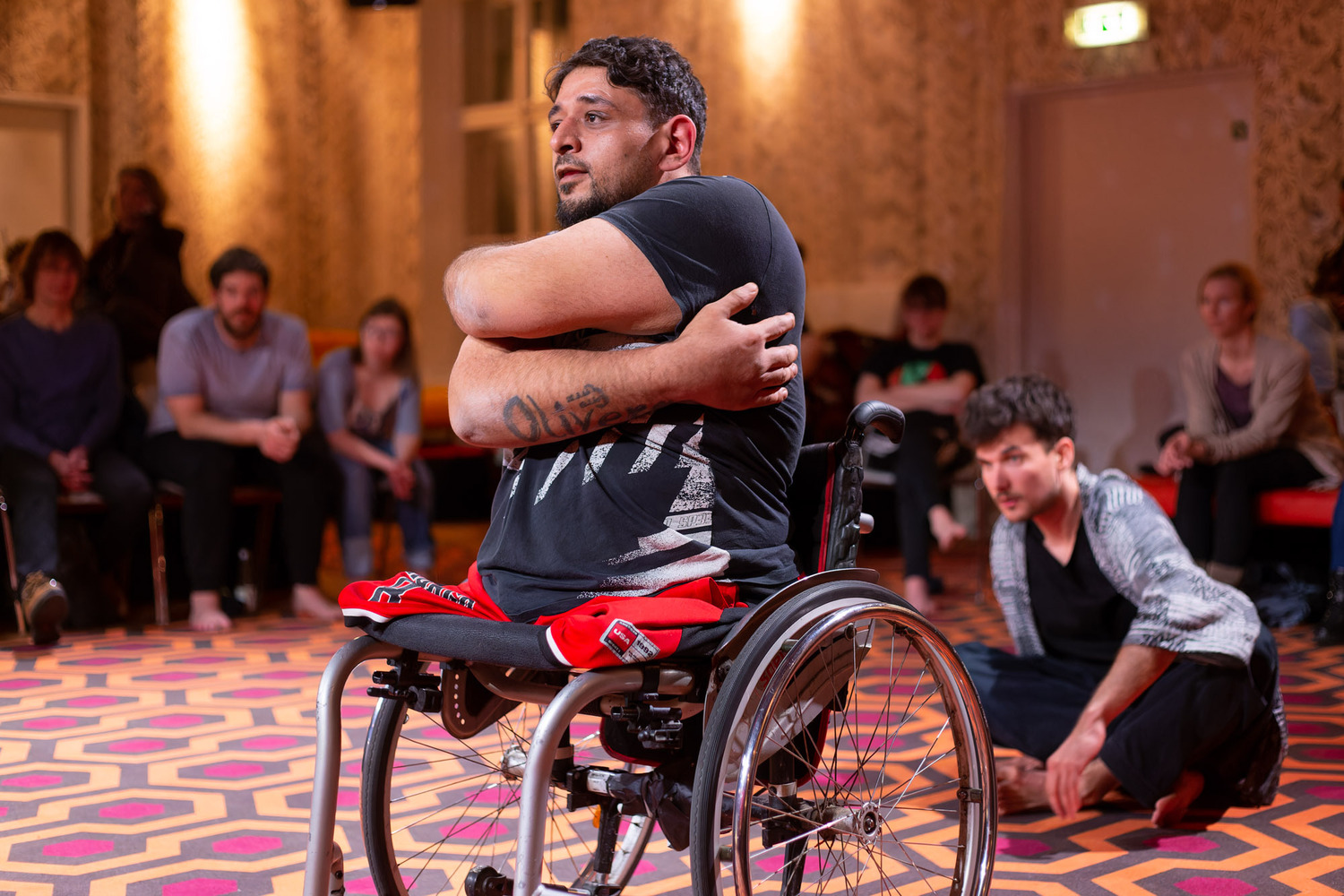 Man sieht einen Raum mit Zuschauer:innen, in der Mitte ein Performer in einem Rollstuhl.