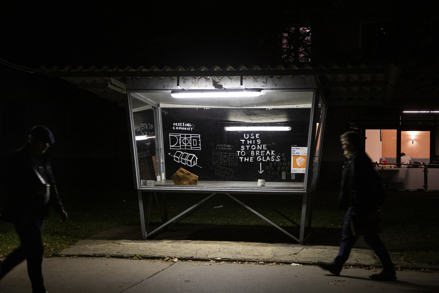 Man sieht eine nächtliche Straßensituation. Zwei Menschen gehen links und rechts an einer Installation im öffentlichen Raum vorbei, einem erleuchteten Kasten, in dem sich ein Stein befinden sowie die Aufforderung, diese zu nutzen, um das Glas zu brechen.