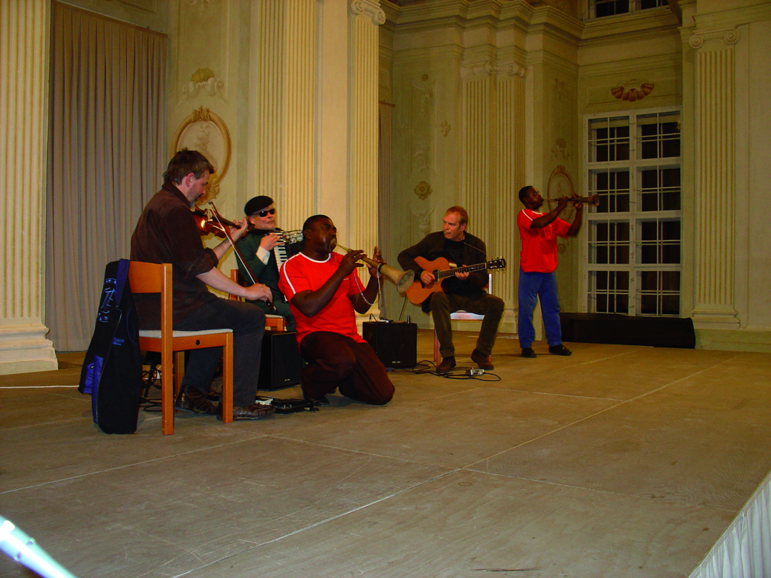 Ein barock wirkender Festsaal/Ballsaal, Säulen, hohe Fenster, goldene Verzierungen, darin spielt eine Gruppe Musiker:innen, Violine, Gitarre, Ziehharmonika, Flöten