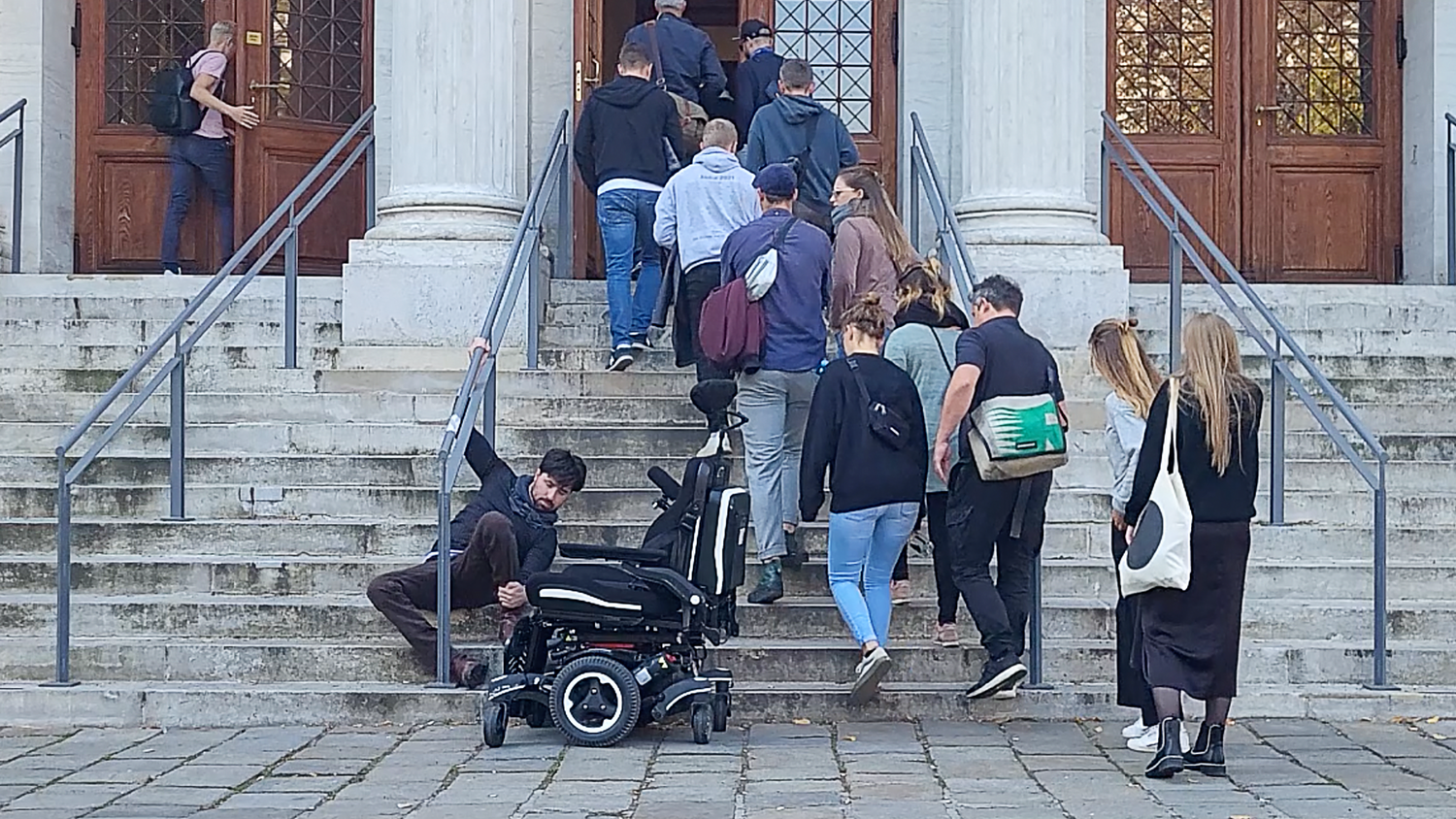 Man sieht mehrere Menschen die Treppen zur Akademie der bildenden Künste hinaufgehen, während eine Person versucht, sich vom Rollstuhl aus die Treppen hinauf zu hanteln.