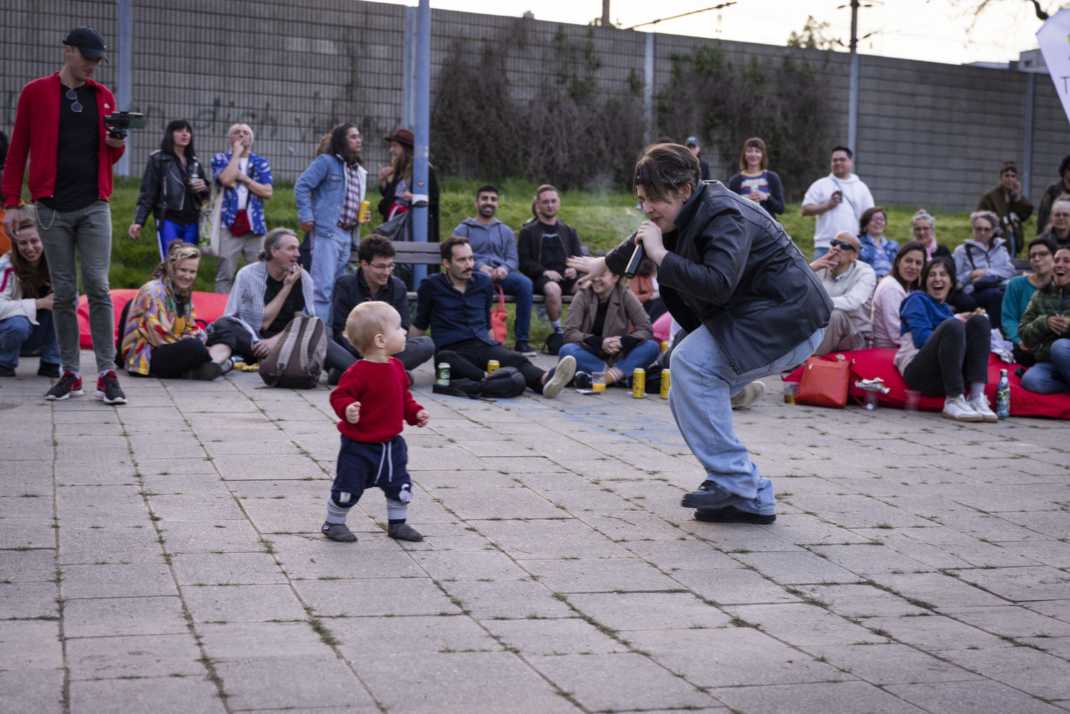 Eine Person singt auf einen offenen Platz in ein Mikro. Um sie sitzen Menschen, vor ihr steht ein Kleinkind und tanzt mit.