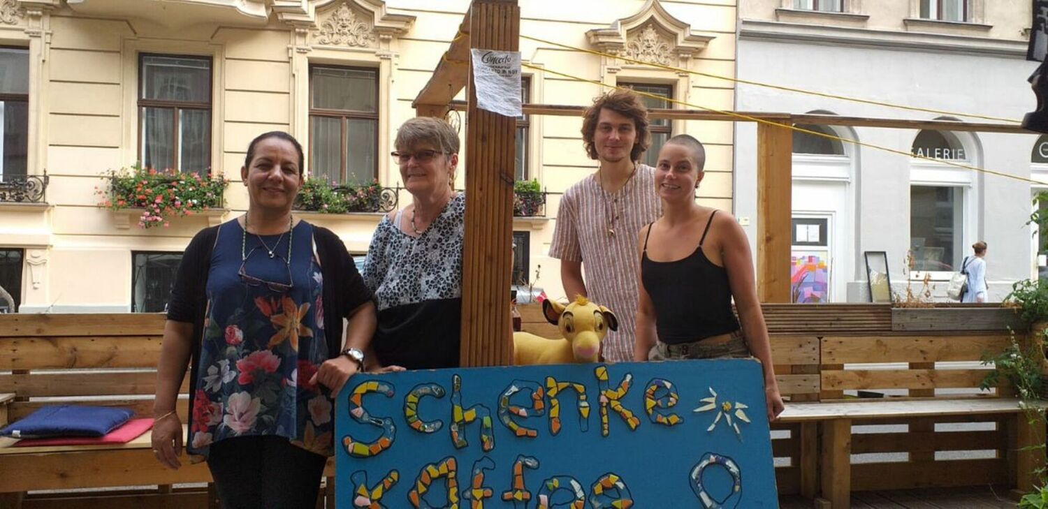 Das Team der Schenke in einem Schanigarten vor einem Plakat des "Schenke-Kaffees"