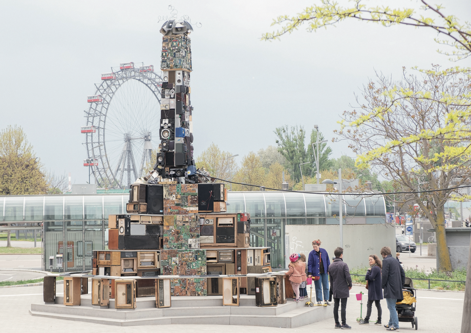 Eine Skulptur im öffentlichen Raum, im Hintergrund das Riesenrad, neben dem Objekt mehrere Menschen im Gespräch.