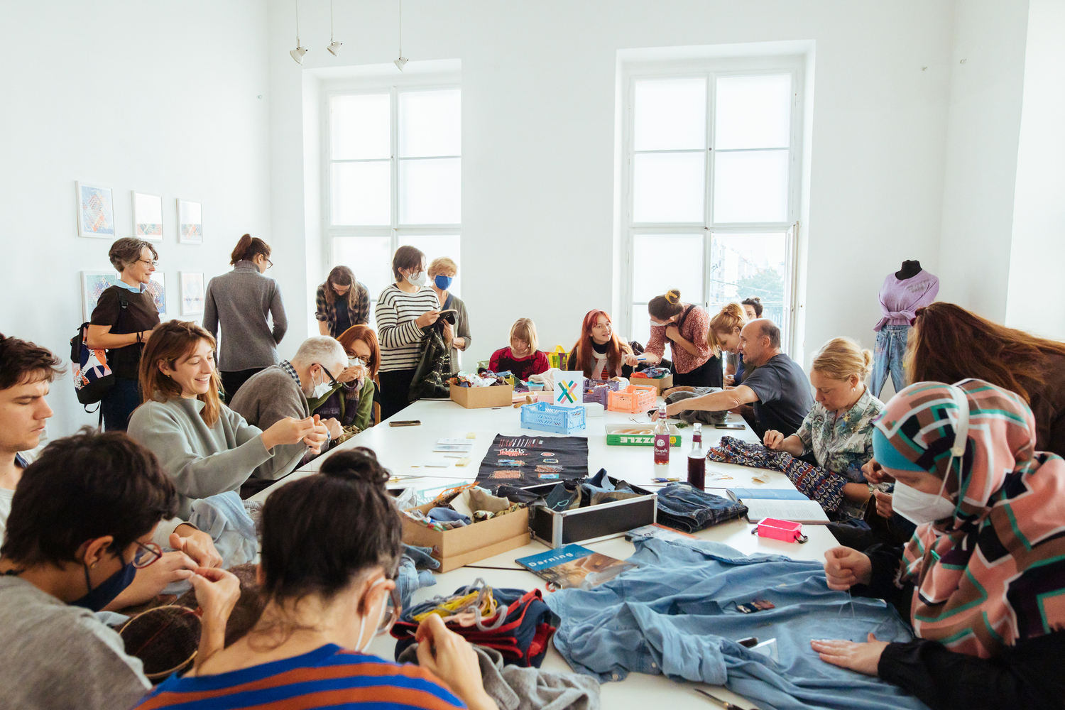 Man sieht viele Personen an einem großen Tisch sitzen und nähen, flicken, plaudern und lachen.