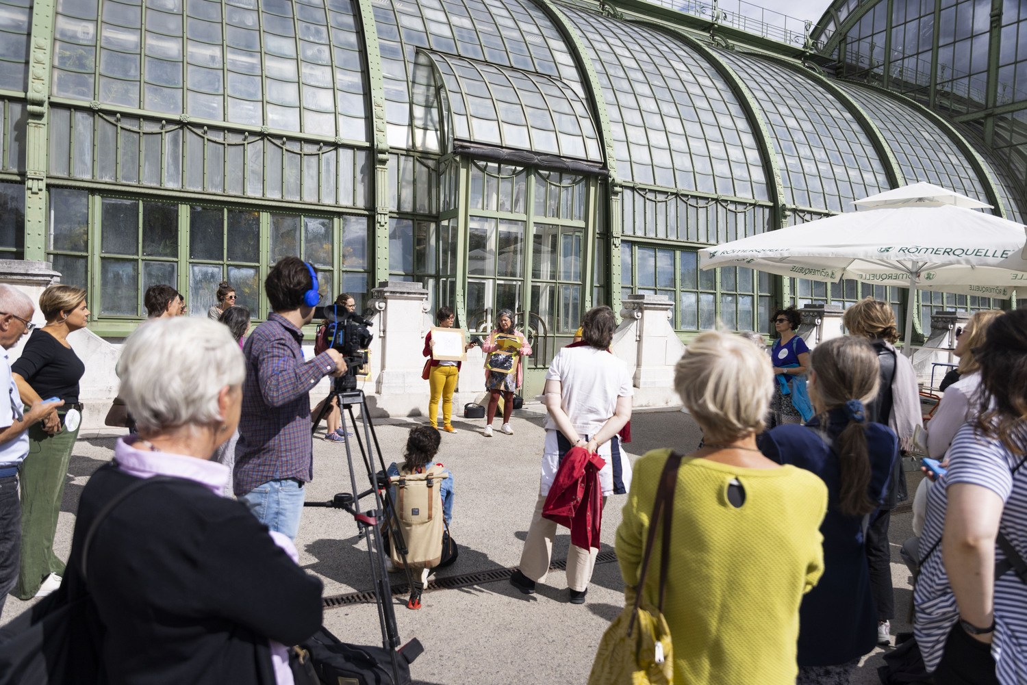 Personen stehen vor dem Palmenhaus in Wien und hören und sehen zwei Vortragenden zu.