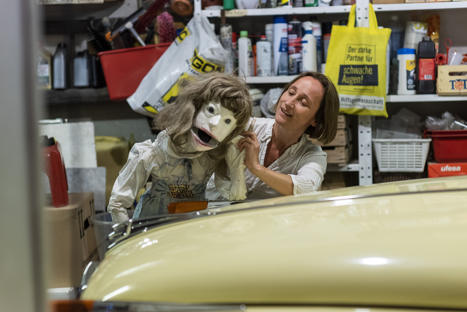 Eine Puppenspielerin und eine Puppe lehnen an einem hellen Auto, das in einer vollgeräumten Garage steht.