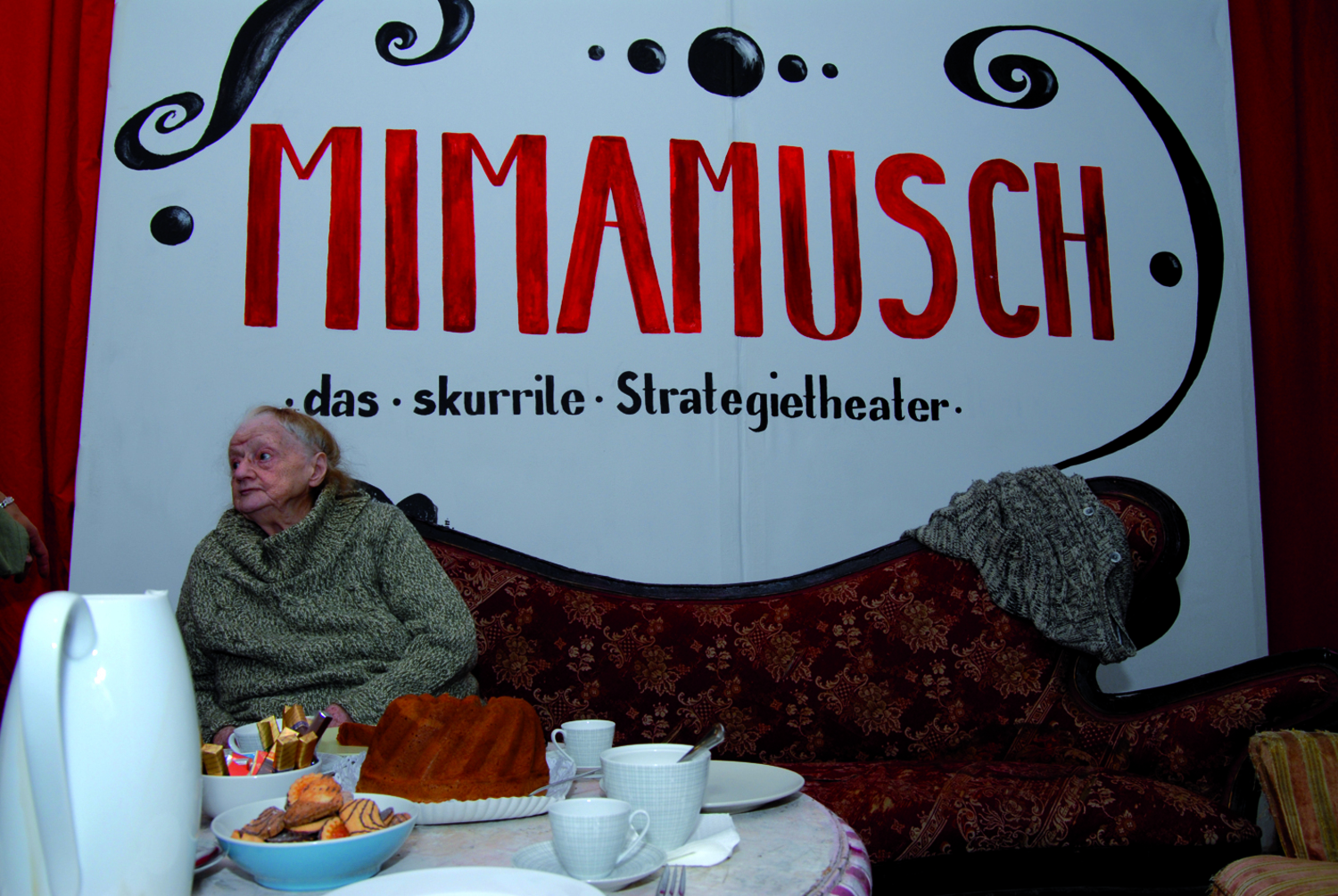 Eine alte Frau sitzt auf einem roten Sofa, davor ein Tisch mit Jause, dahinter ein großes Plakat an der Wand mit handschriftlichem Titel/Werbetext für Mimamusch